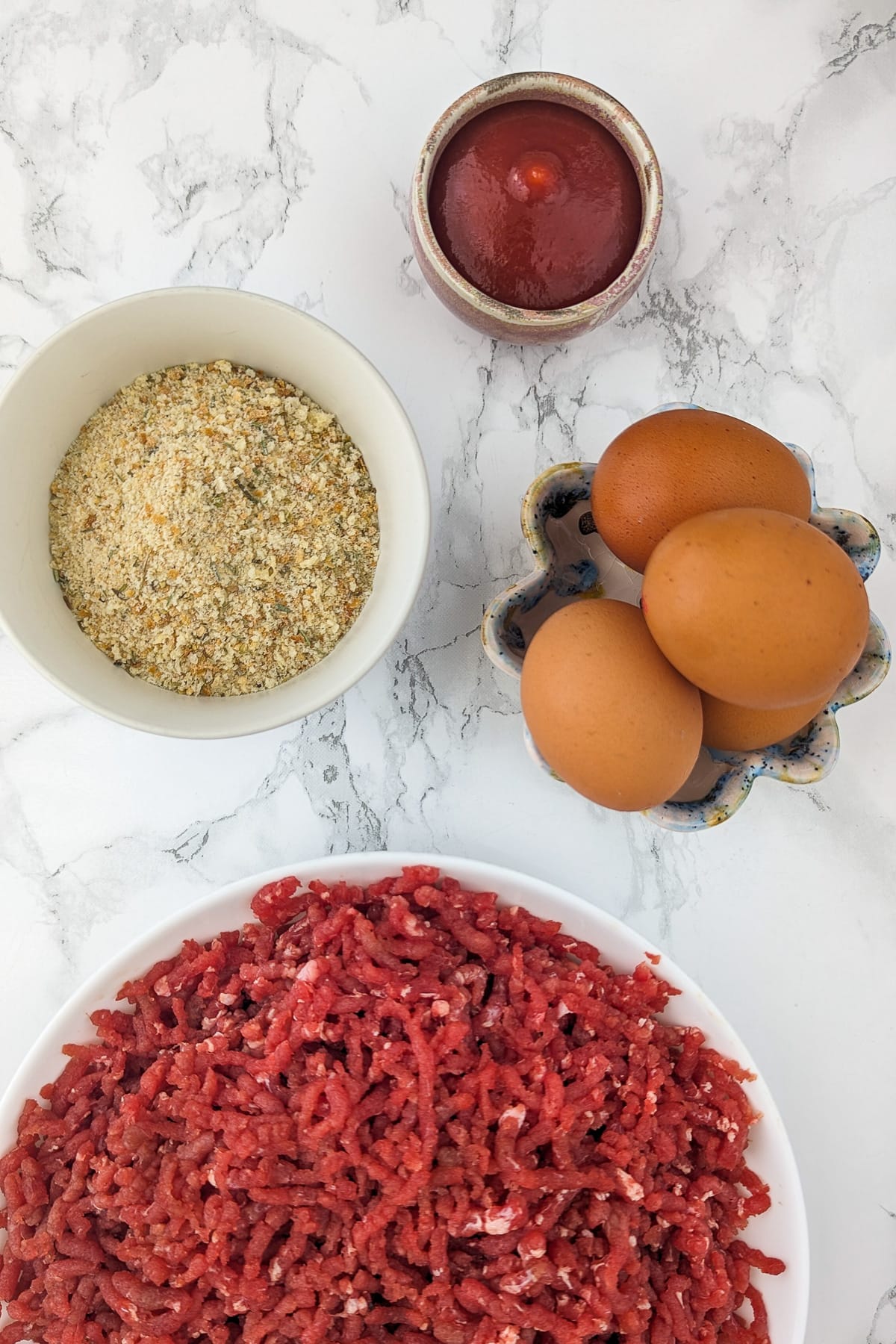 Top view of a white bowl with ground beef, 4 eggs, ketchup, breadcrumbs, and spices.