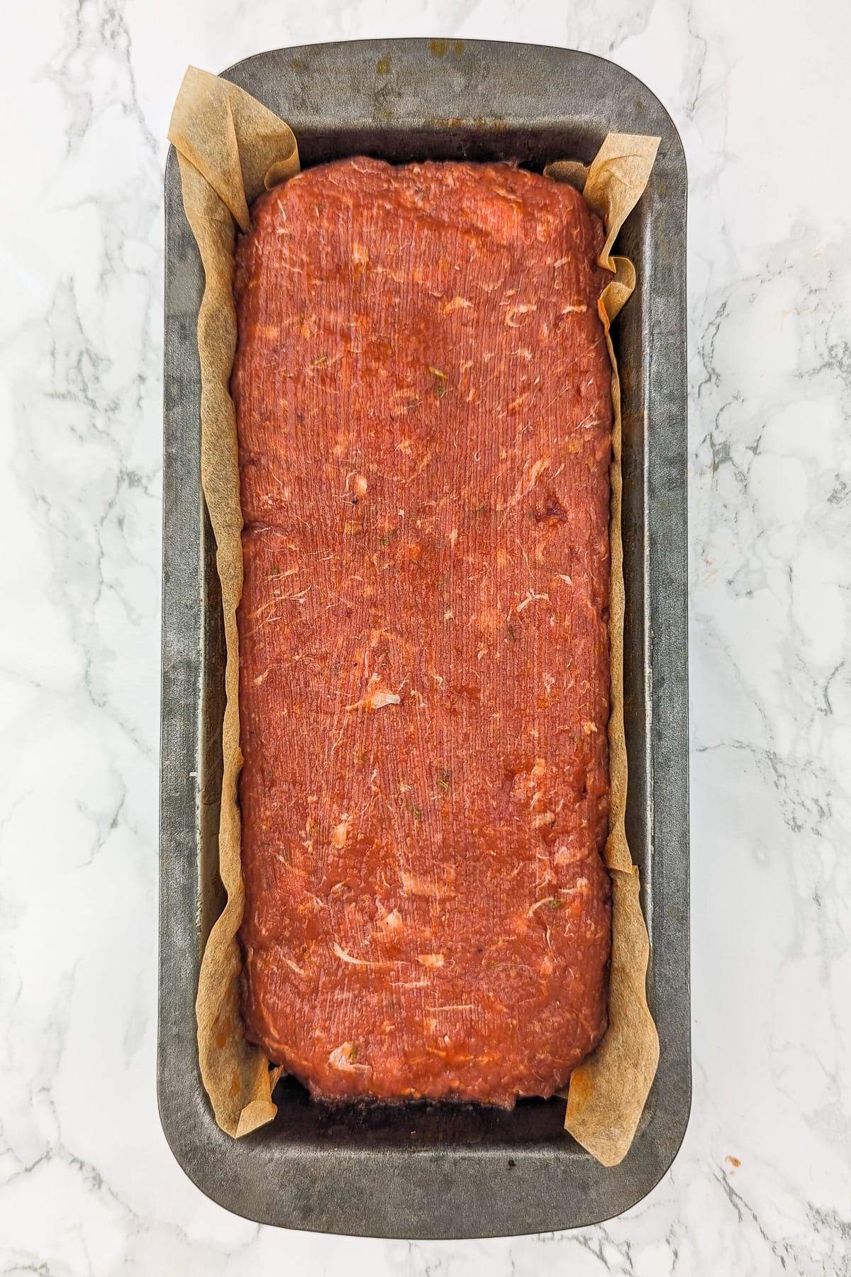 Top look of a raw meatloaf in a metal baking tray sitting on a white marble table.