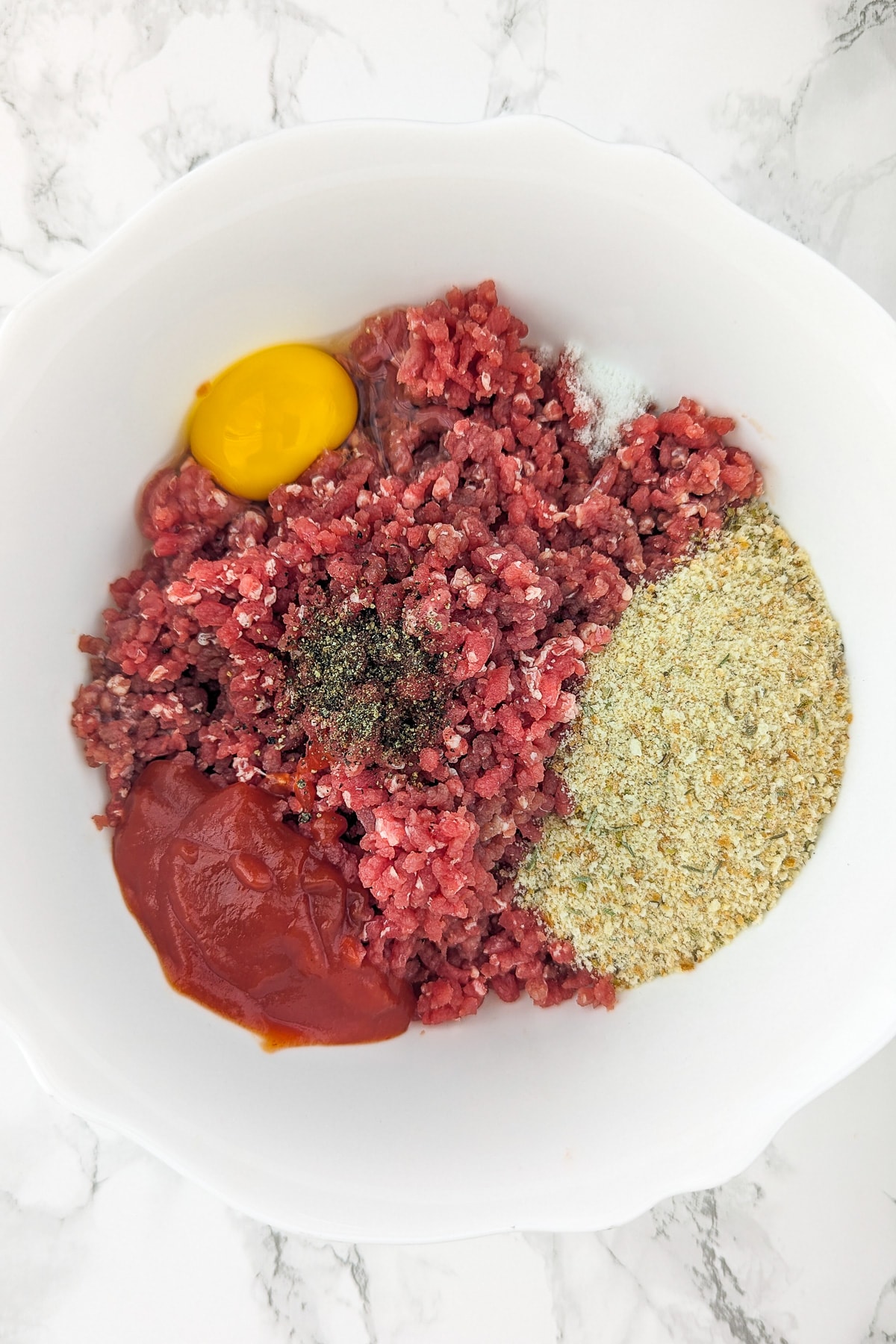 Top view of a white bowl with ground beef, egg, ketchup, breadcrumbs, and spices.