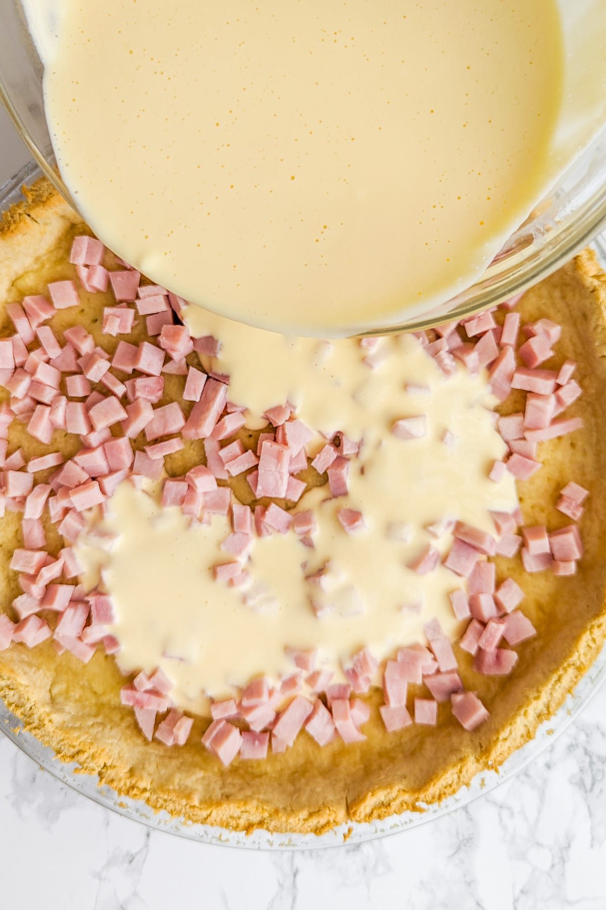 Pouring egg and cream cheese mix over a tart with ham.