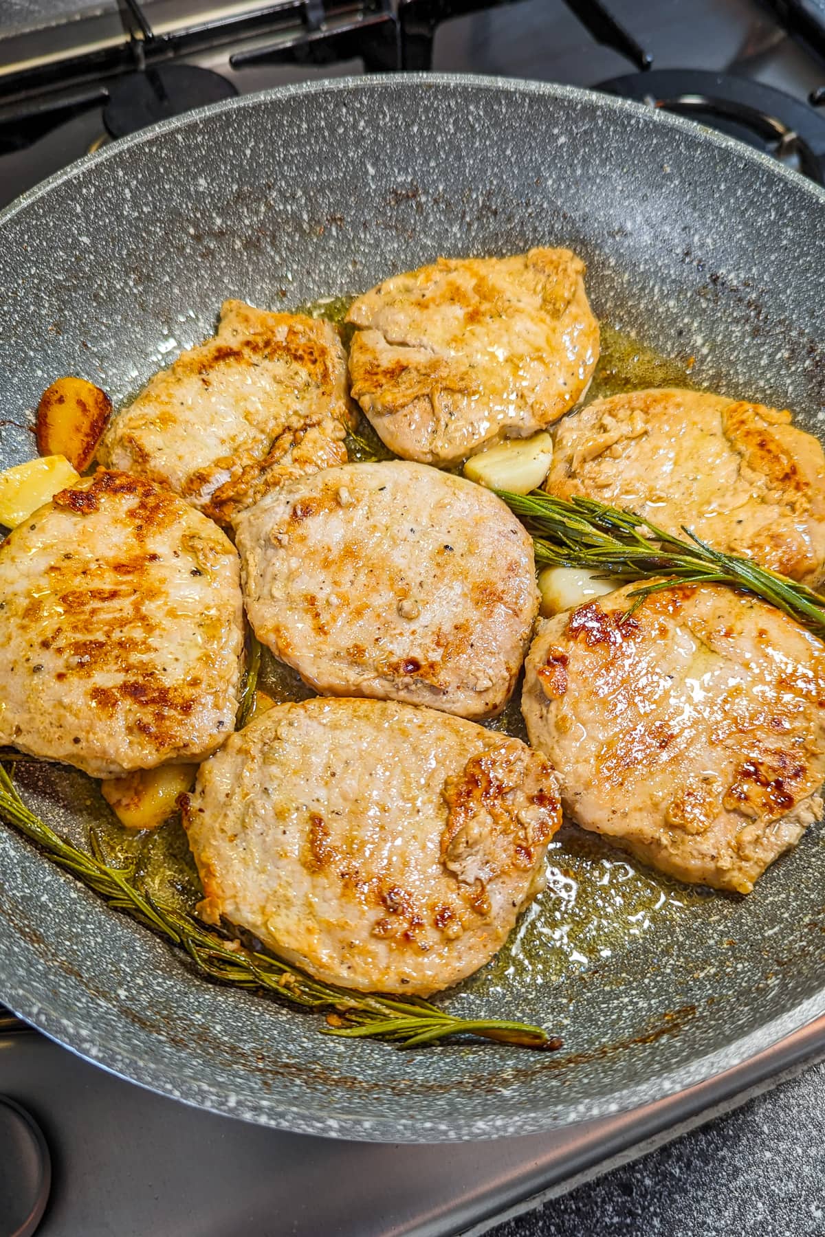 Top view of 7 pork chops in a frying pan with rosemary brunches.