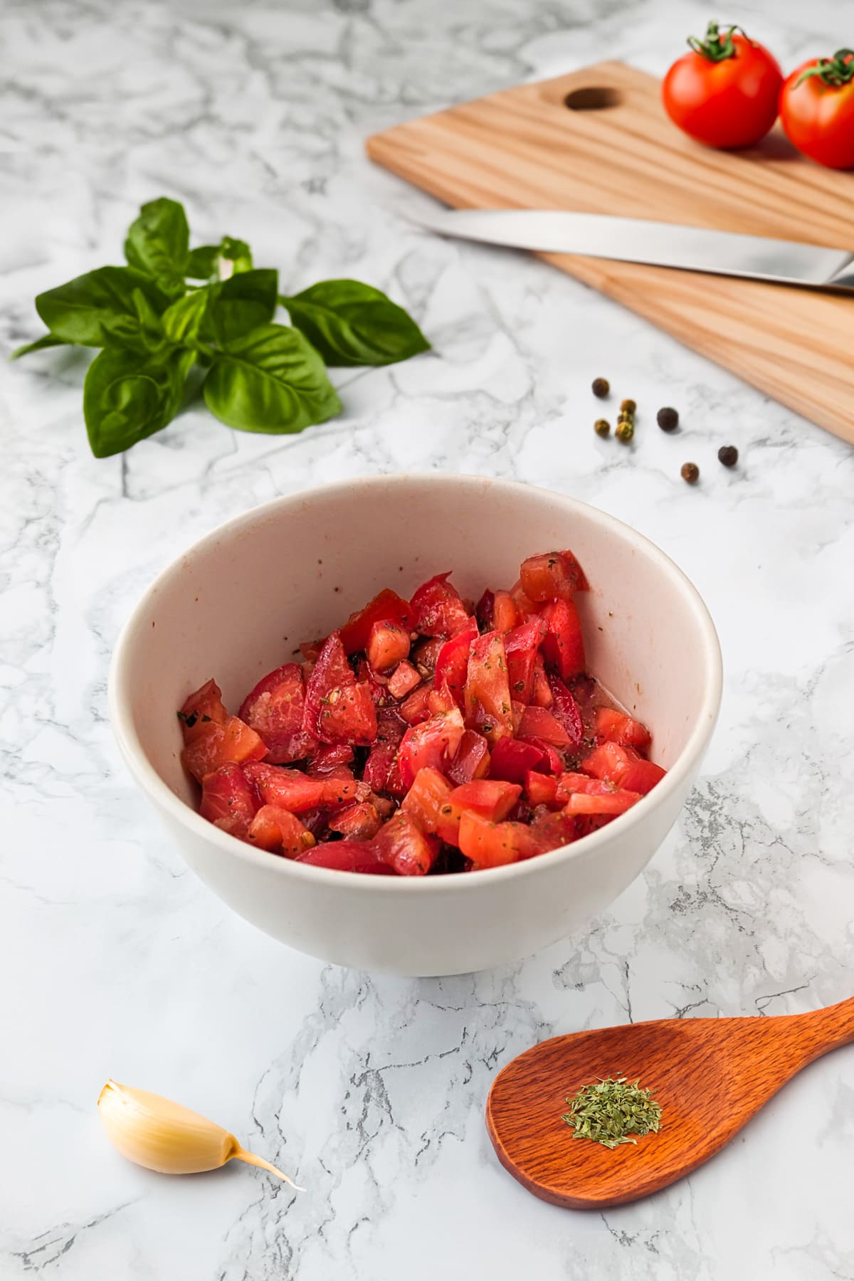 Side view of a white bowl with sliced tomatoes near a wooden spoon with dried oregano, garlic and basil.