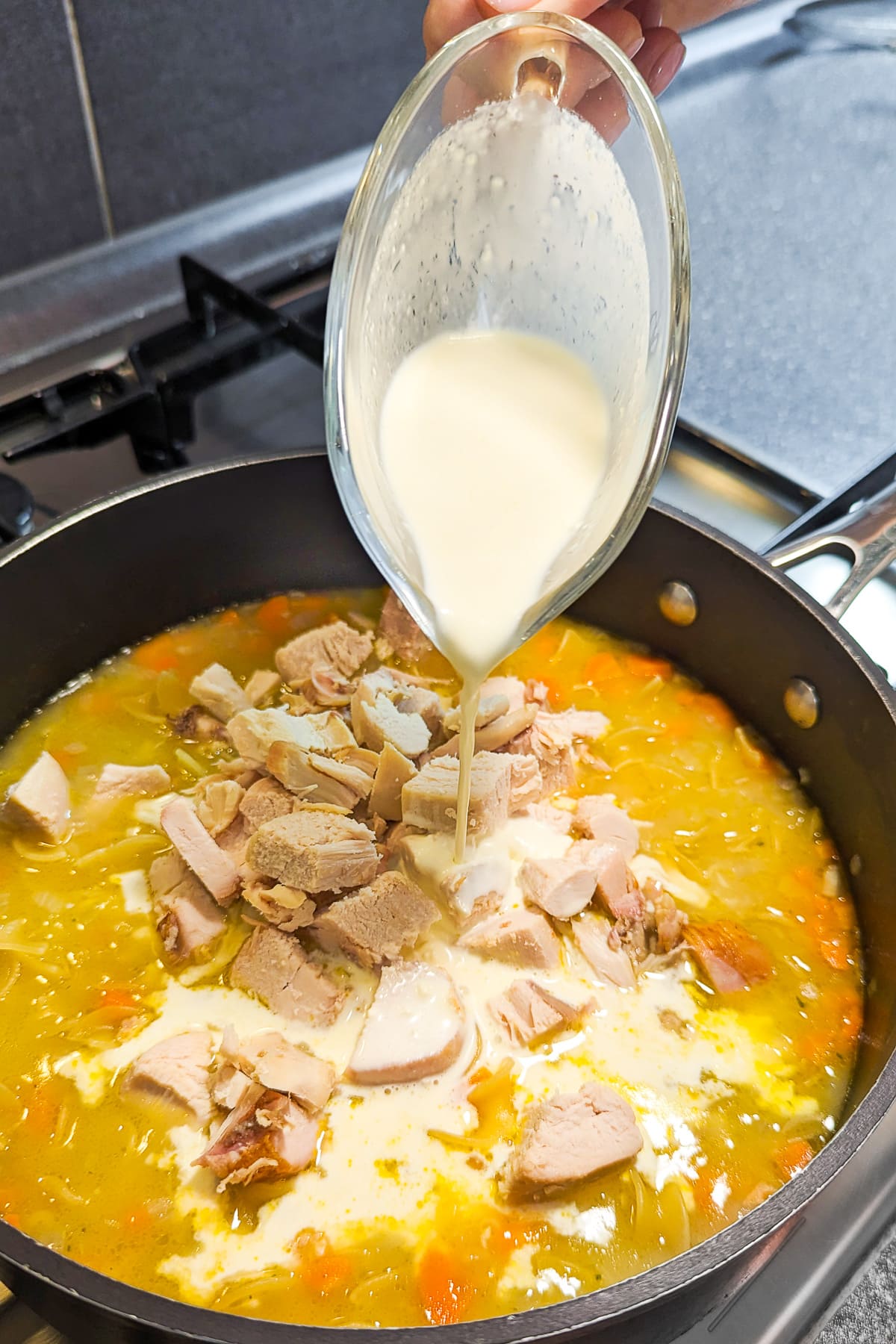 Pouring cream over turkey meat and vegetable soup in a deep frying pan on the stove.