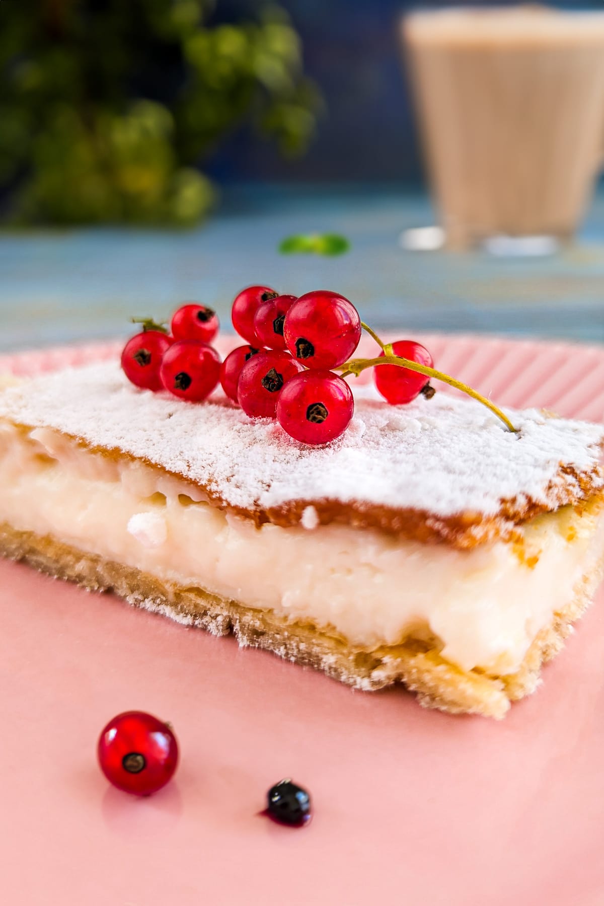 Close look of a vanilla slice on a pink plate with a cappucino in the background.