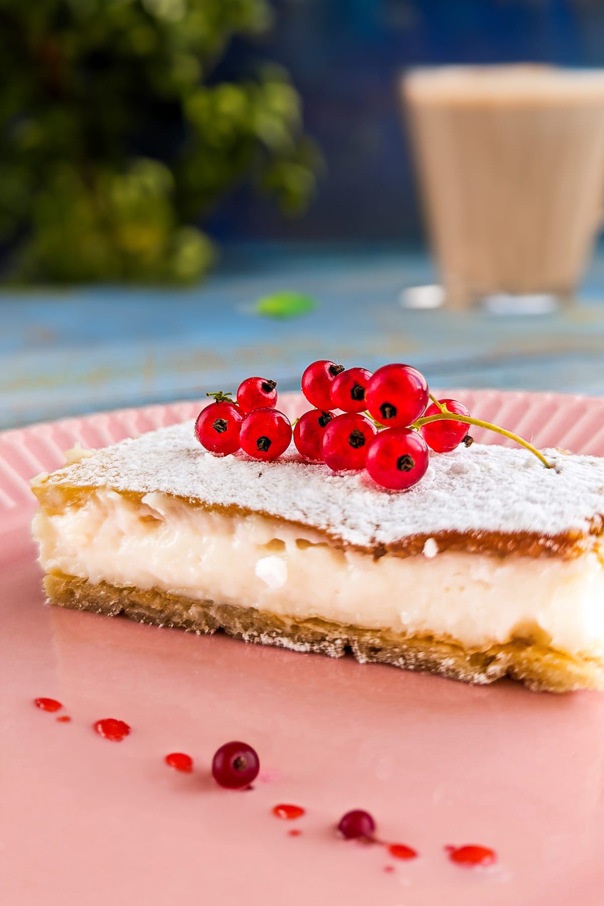 Close look of a vanilla slice on a pink plate with a cappucino in the background.