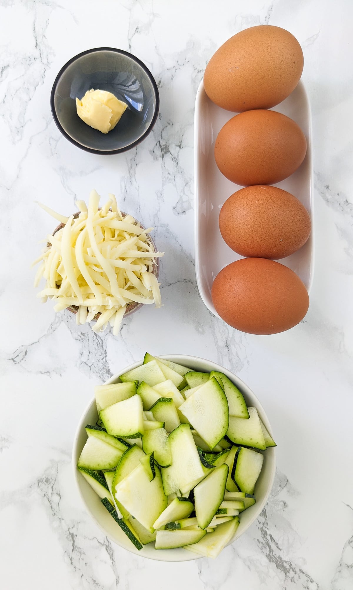 4 eggs, sliced zucchini, mozzarella cheese and butter on a white marble table.
