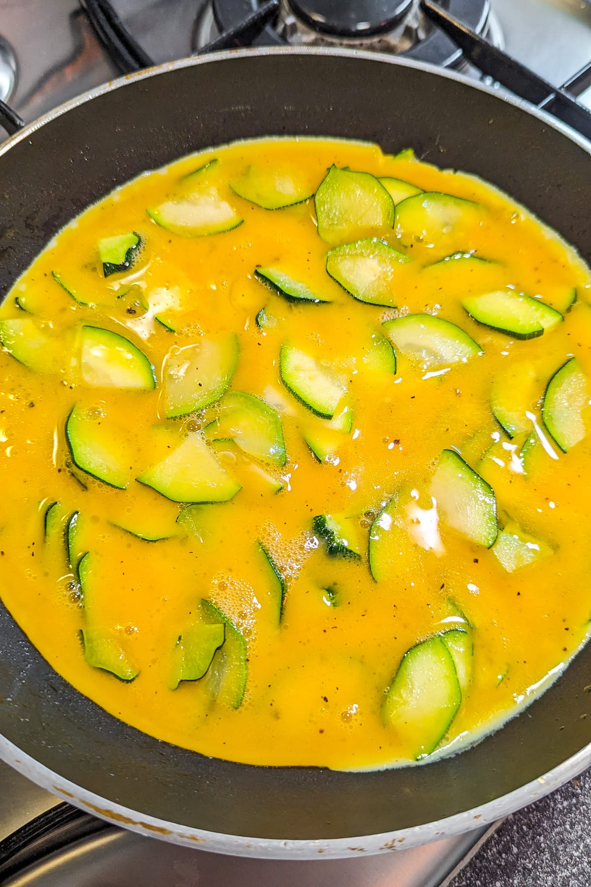 Sliced zucchini covered with beaten eggs in a frying pan on the stove.