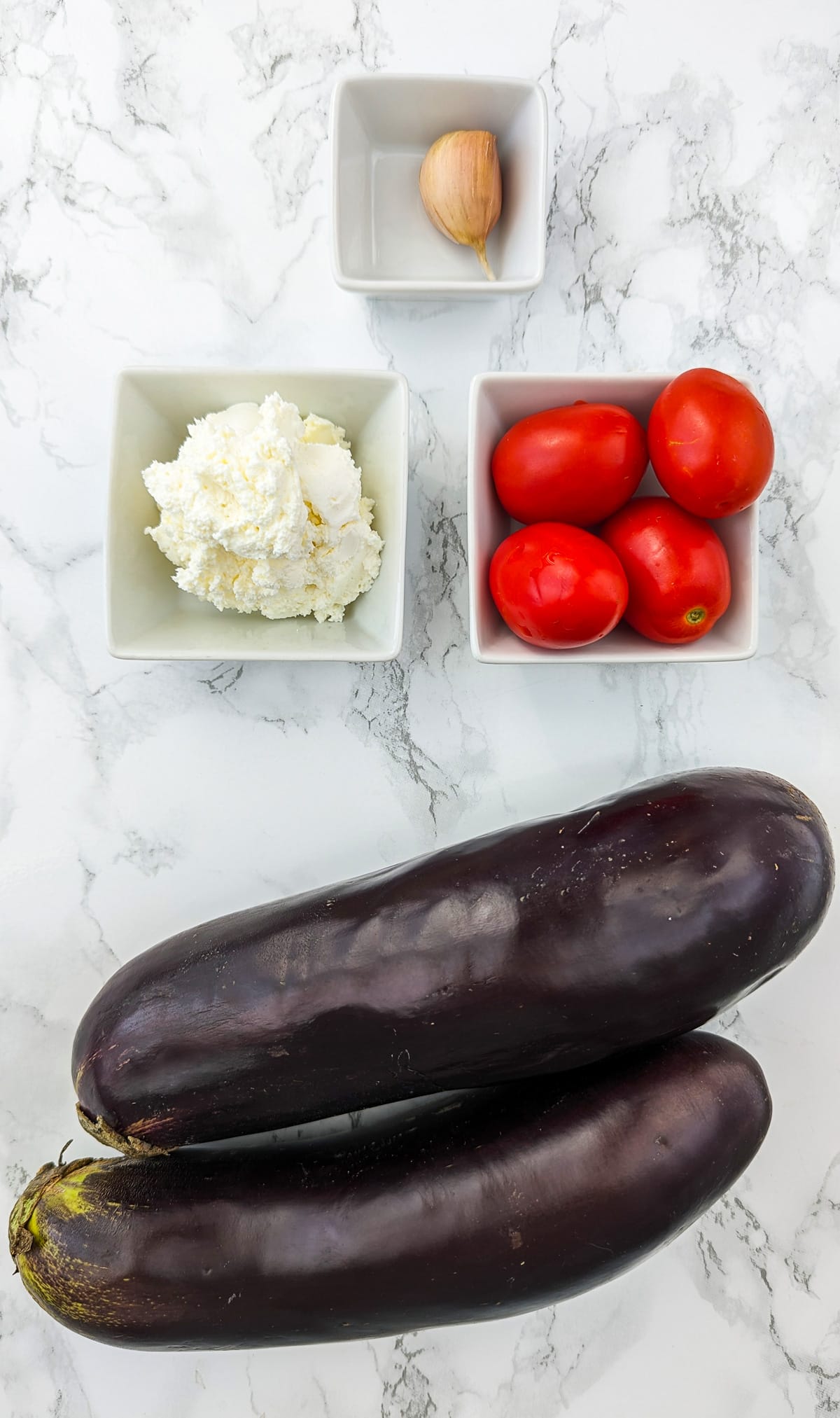 Top view of 2 large eggplants near 4 tomatoes, cream cheese and garlic clove.