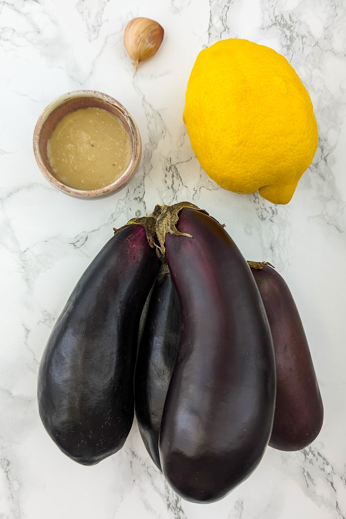 4 eggplants, lemon, tahini and garlic clove on a white marble table.