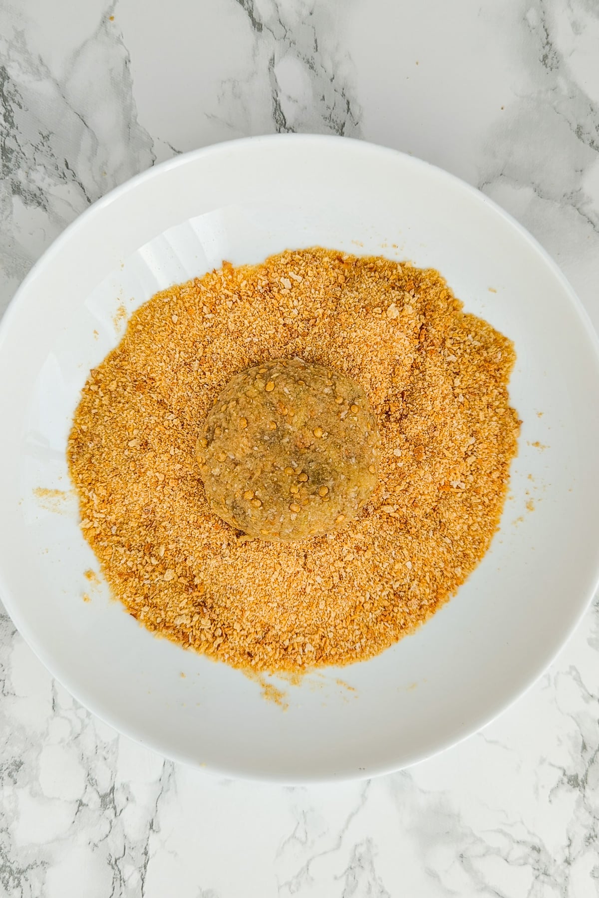 Top view of a white plate with breadcrumbs and eggplant patties coated in breadcrumbs.