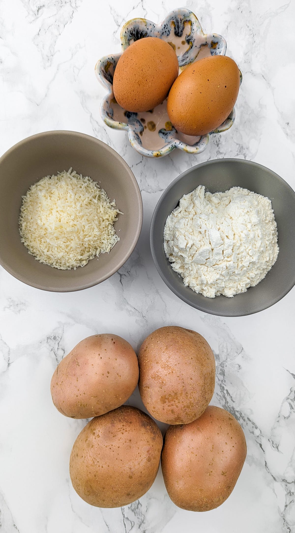 Top view of 4 potatoes, flour, parmesan and 2 eggs on a white marble table.