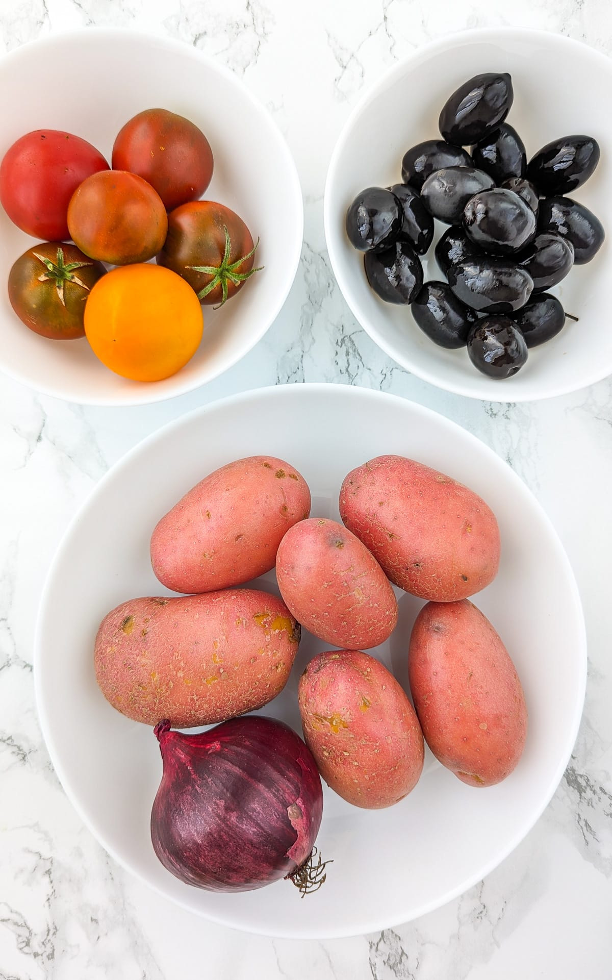 Top view of a plate with red potatoes and onions near a plate of black olives and colorful tomatoes.