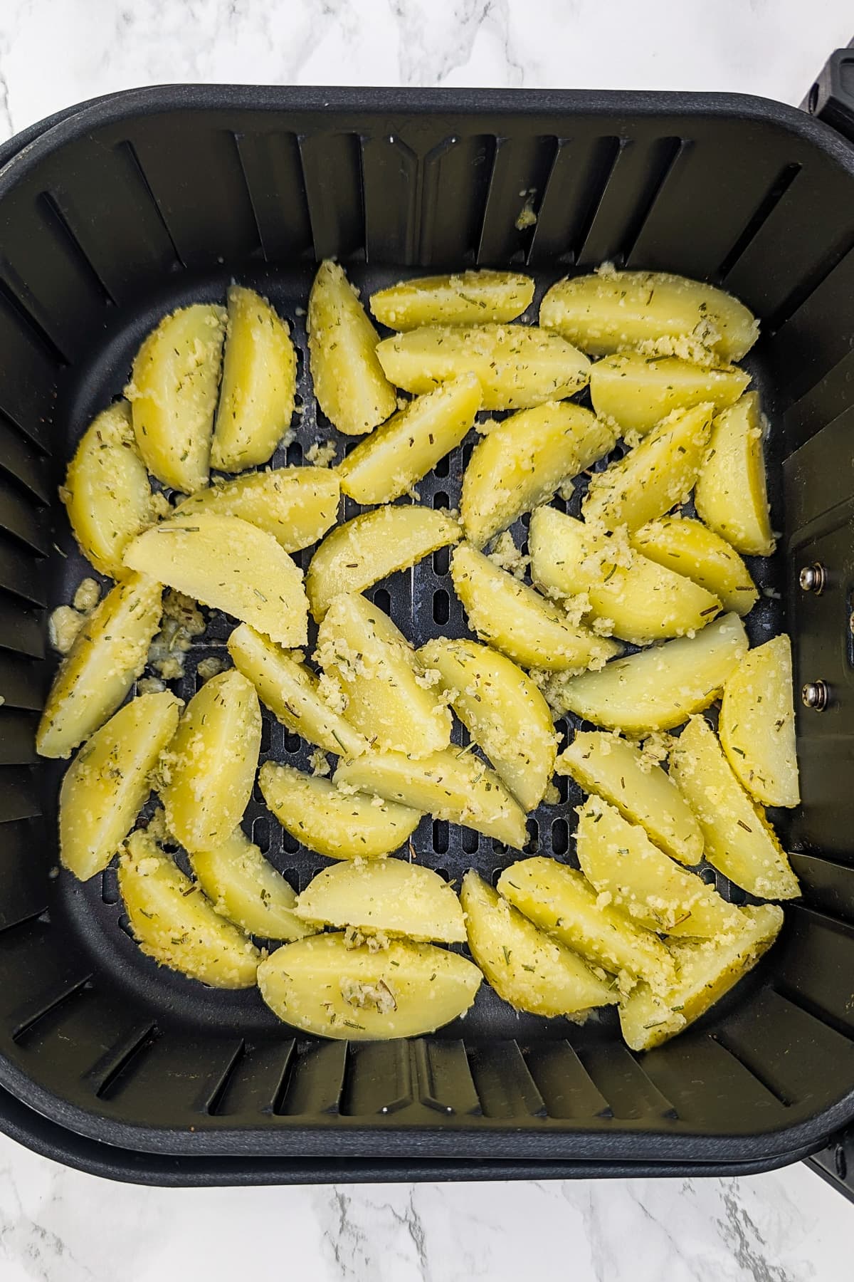 Top view of air fryer basket with potato wedges seasoned with garlic, rosemary and parmesan.