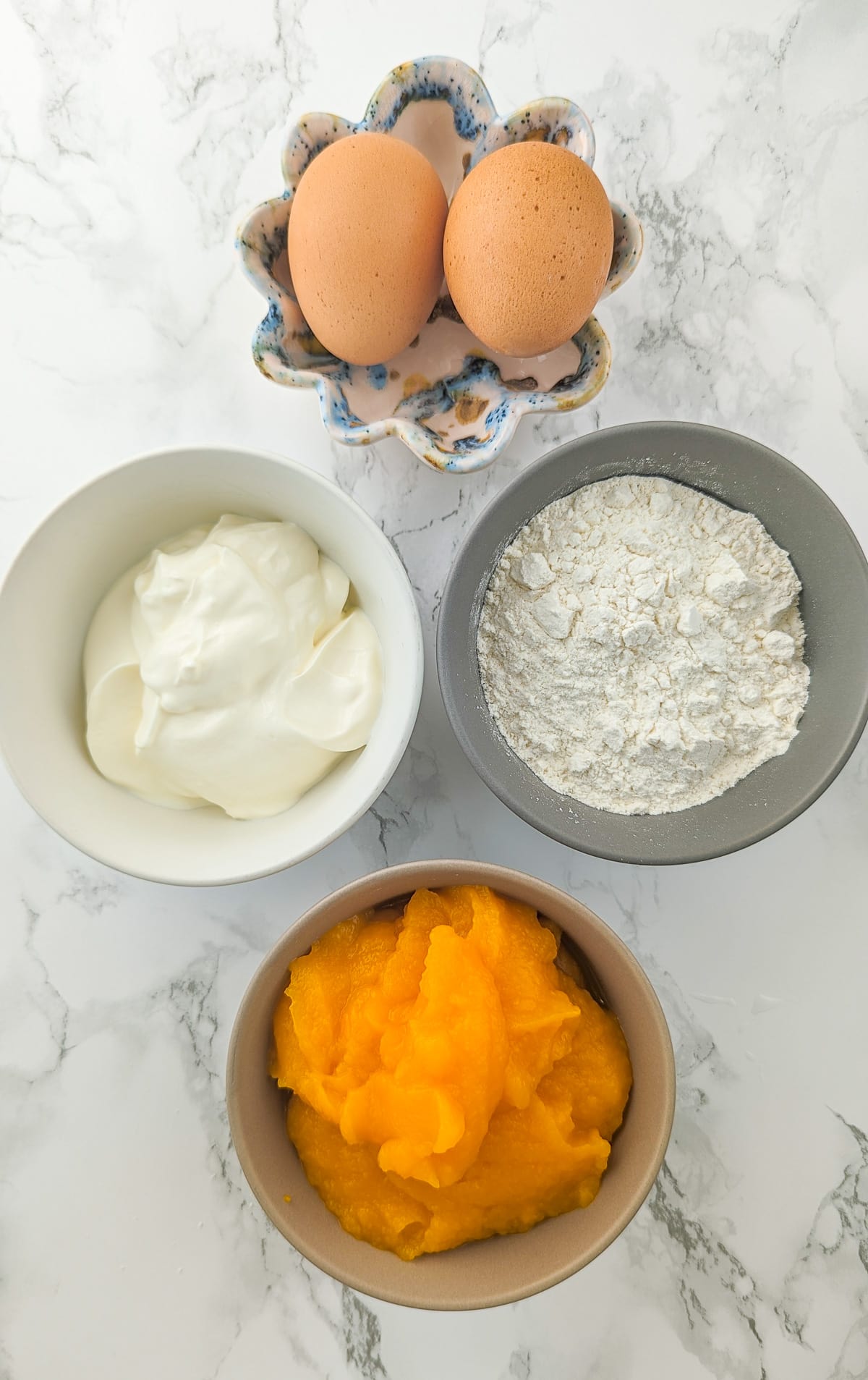 2 eggs, flour, sour cream and pumpkin puree on a white marble table.