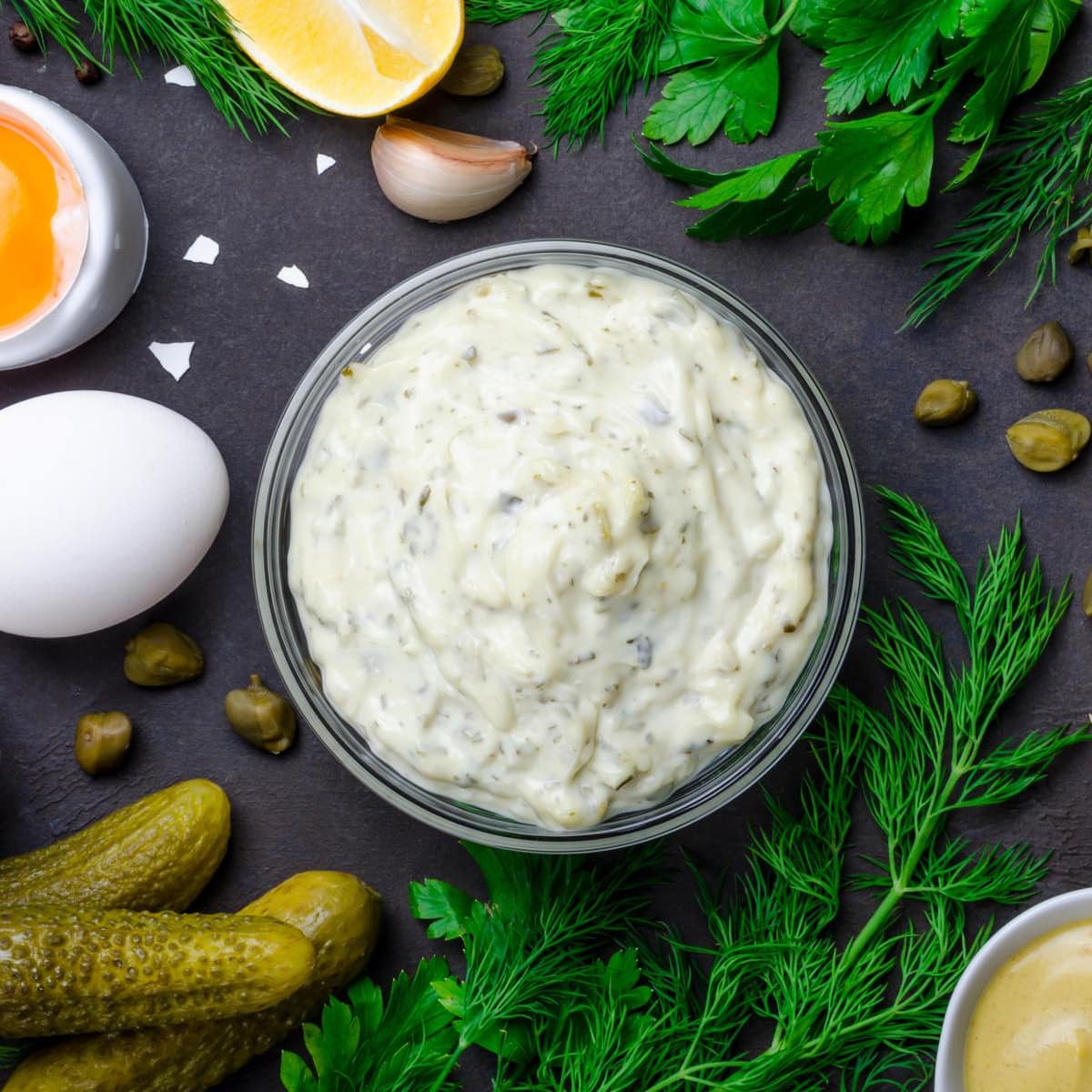 Top view of tartar sauce near dill, cucumbers, garlic, capers and eggs.