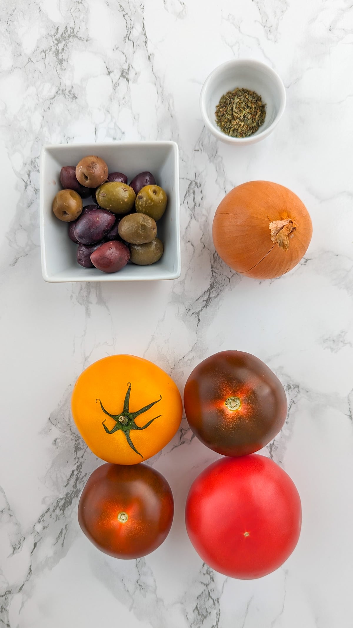 Top view of 4 tomatoes, an onion and olives on a white marble table.
