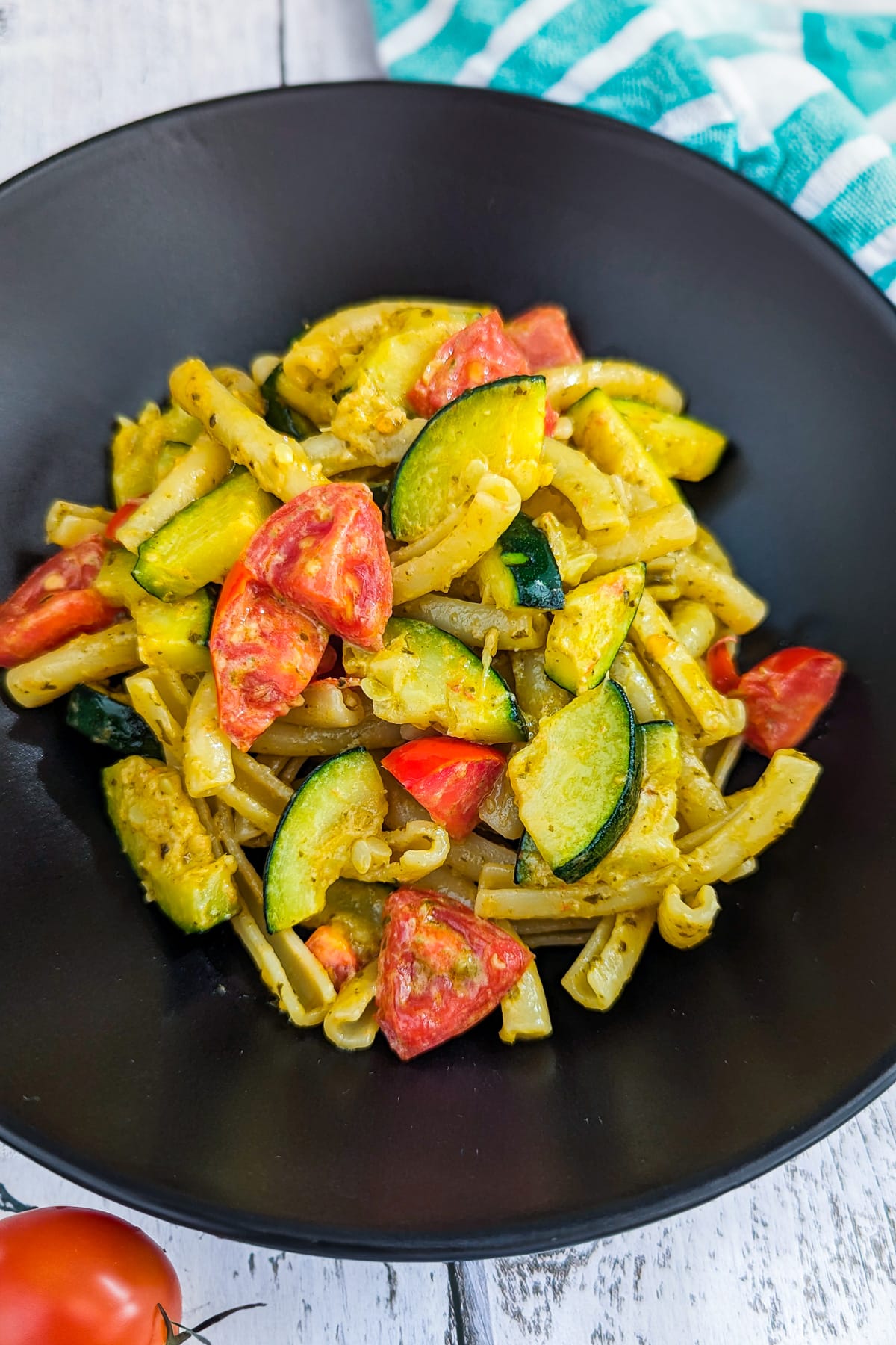 Black plate with zucchini and tomato pasta on a white wooden table.
