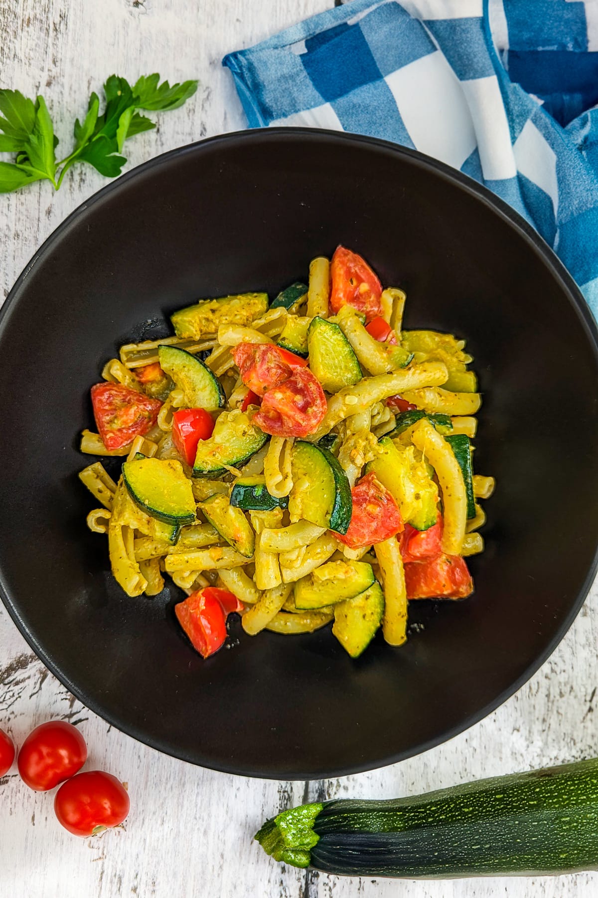 Black plate with zucchini and tomato pasta on a white wooden table.