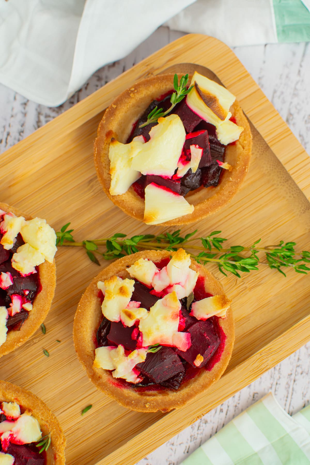 Wooden serving plate with beetroot and feta tartlets near a kitchen towel.