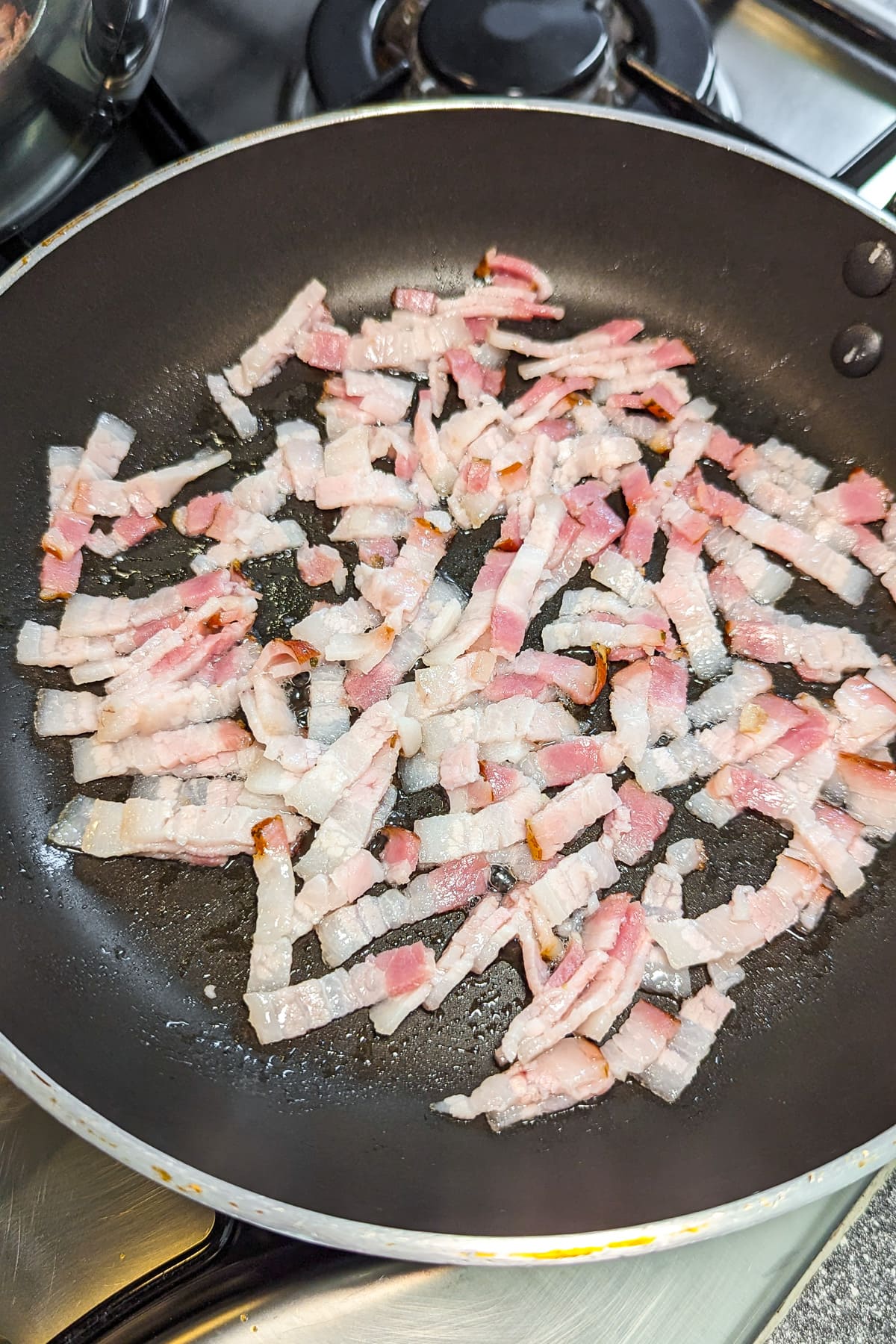 A frying pan with sliced bacon pieces frying on the stove.