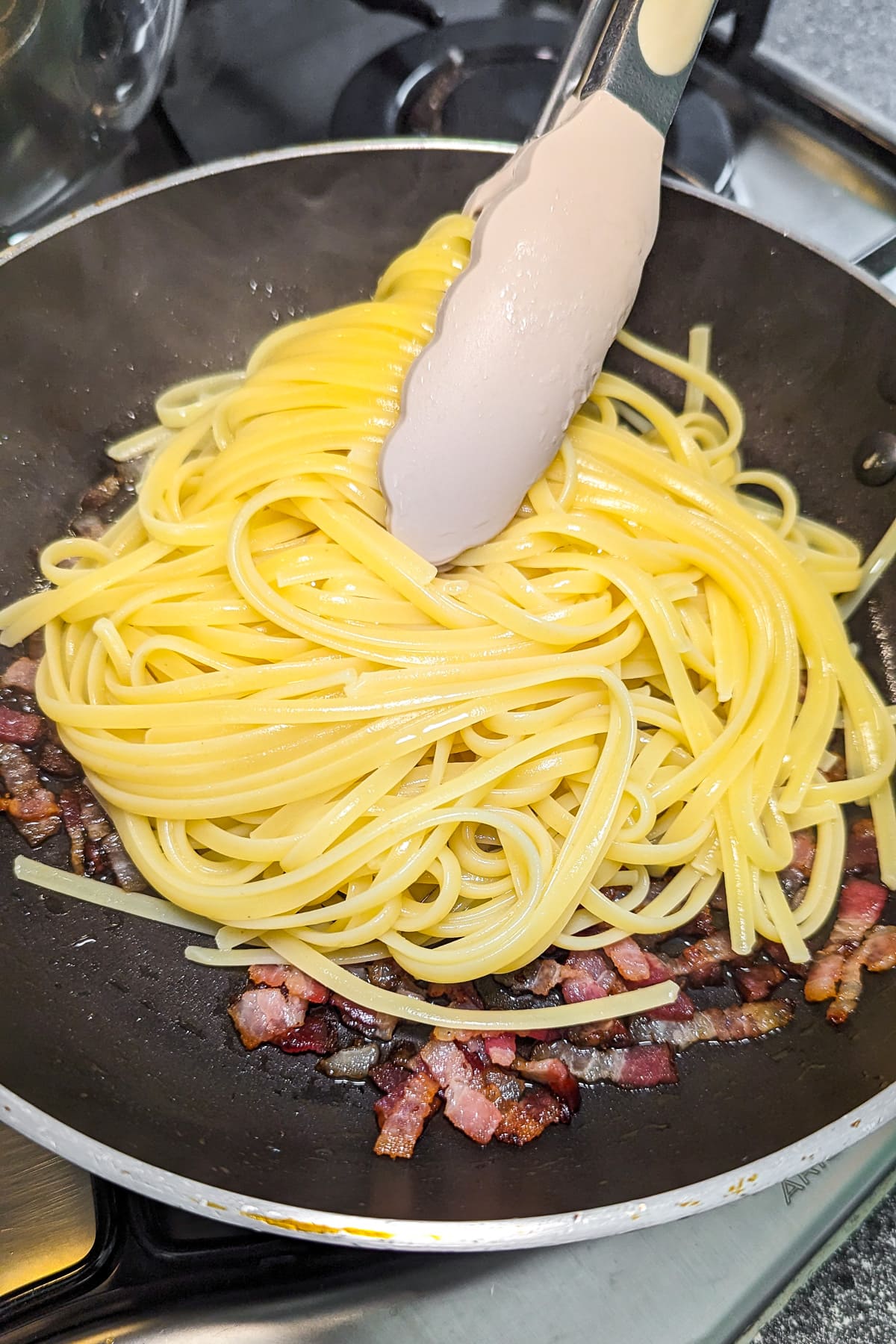 A spatula that holds cooked spaghetti over fried bacon in a frying pan.