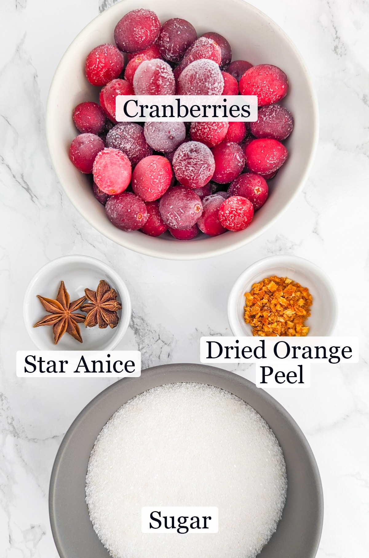 Frozen cranberries, dried orange peel, sugar and star anice on a white marble surface.