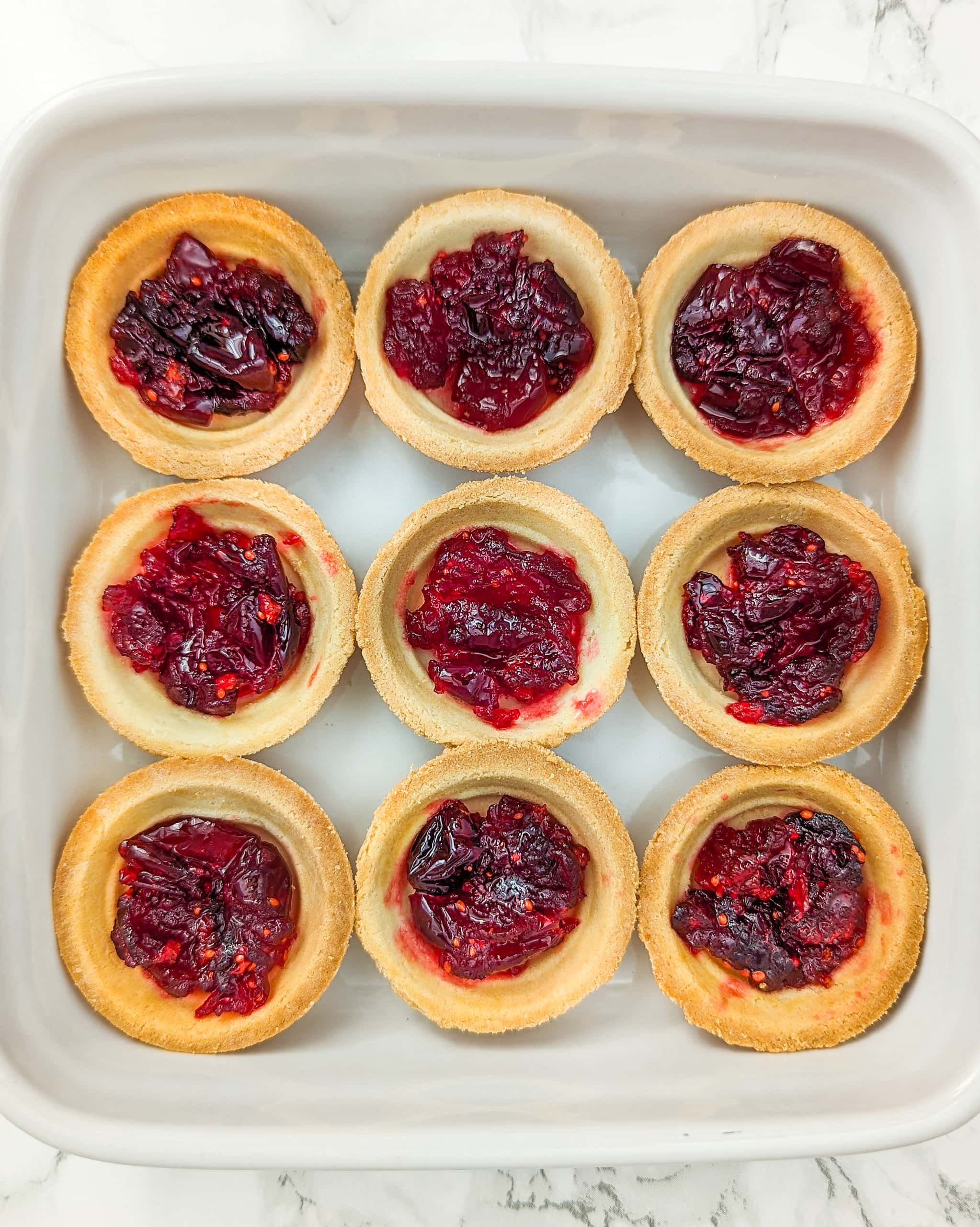 White traybake with cranberry jam in 9 tartlets on a white surface.