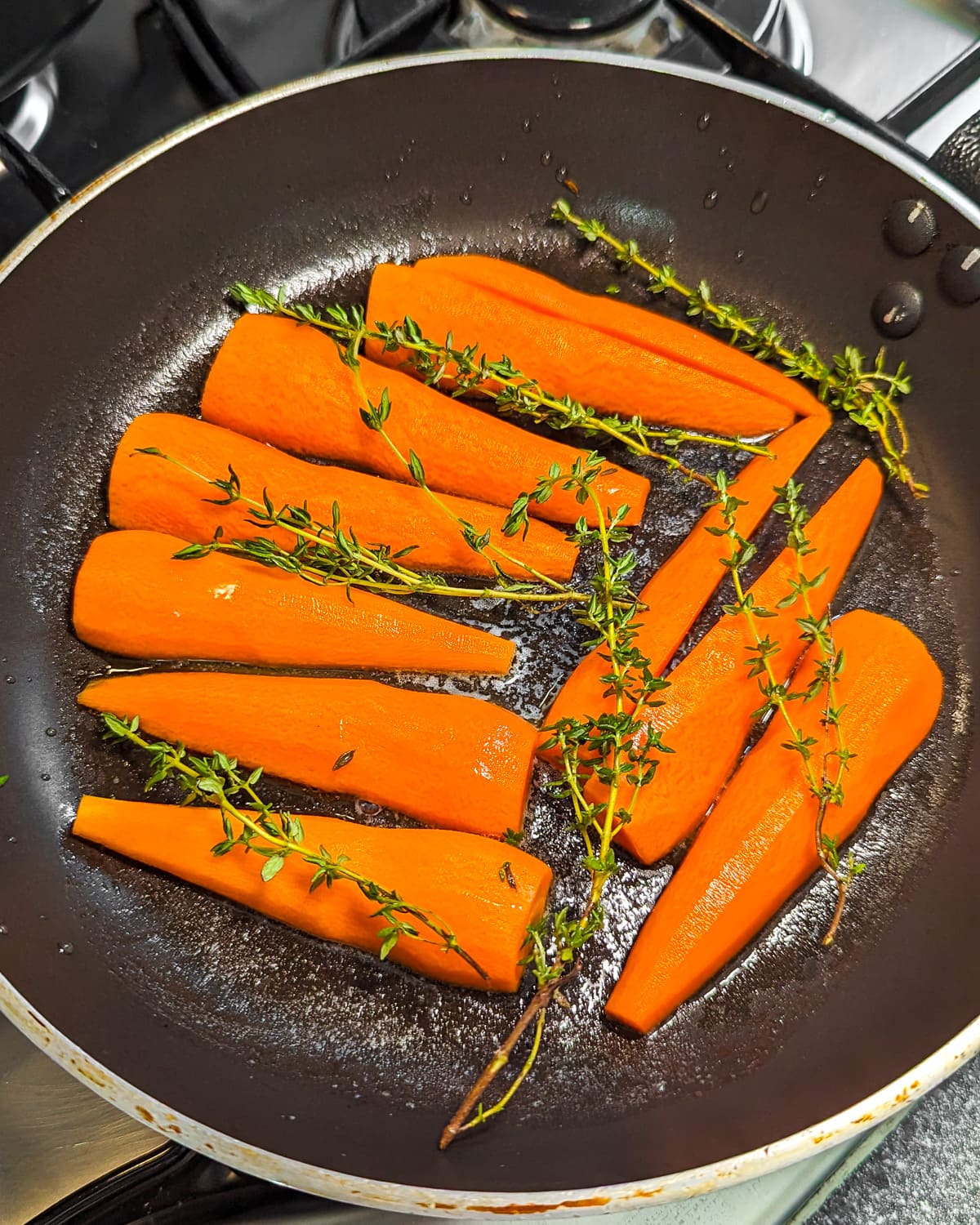 Carrots halves with thyme in a frying pan with melted butter on the stove.