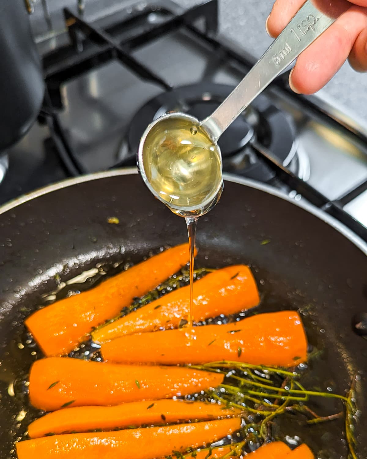A woman hand holding a teaspoon with honey over fried carrots in a frying pan.