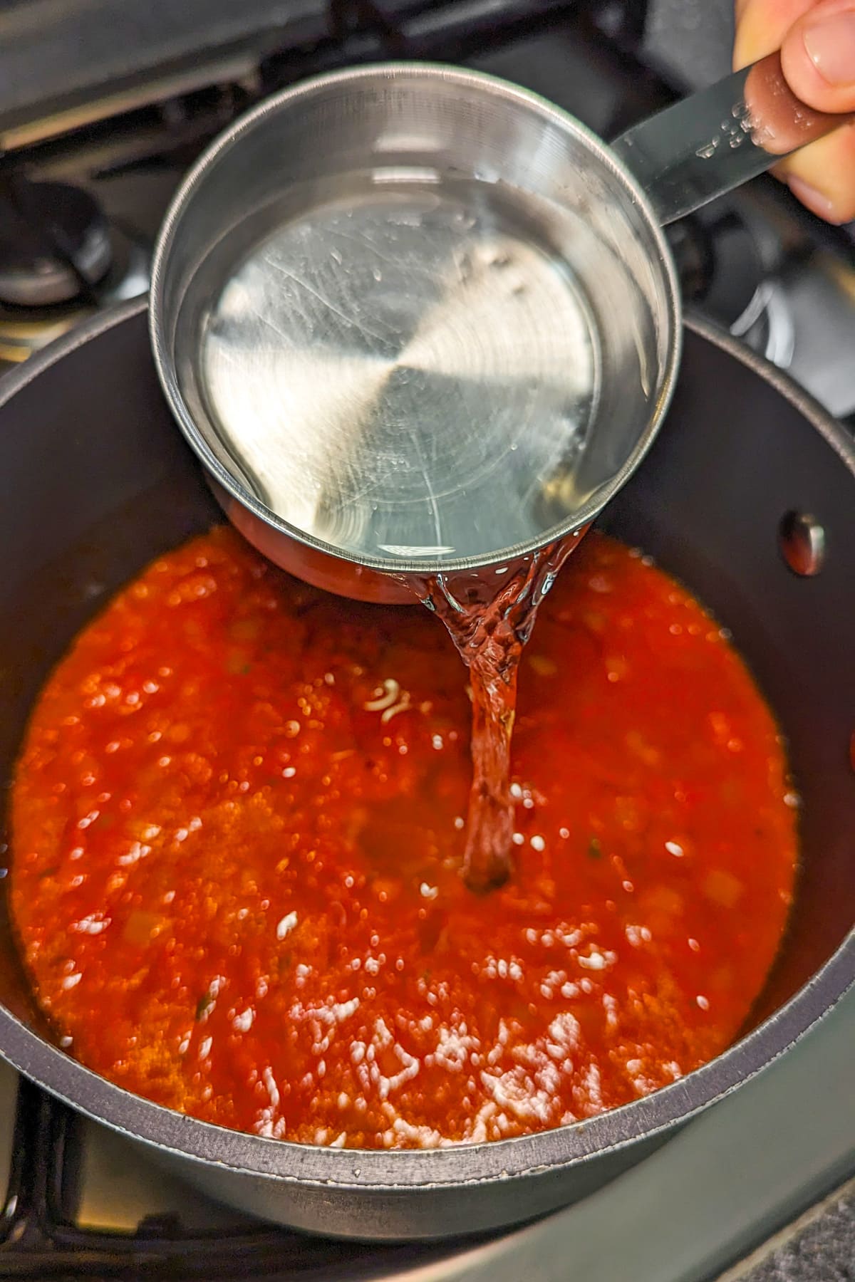 Pouring water over a tomato soup in a pan on the stove.