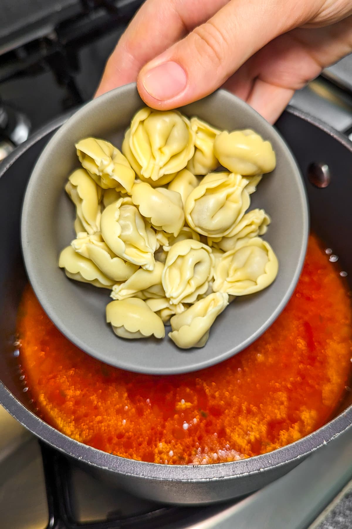 A woman hand holding a bowl with tortellini over a tomato soup in a pan on the stove.