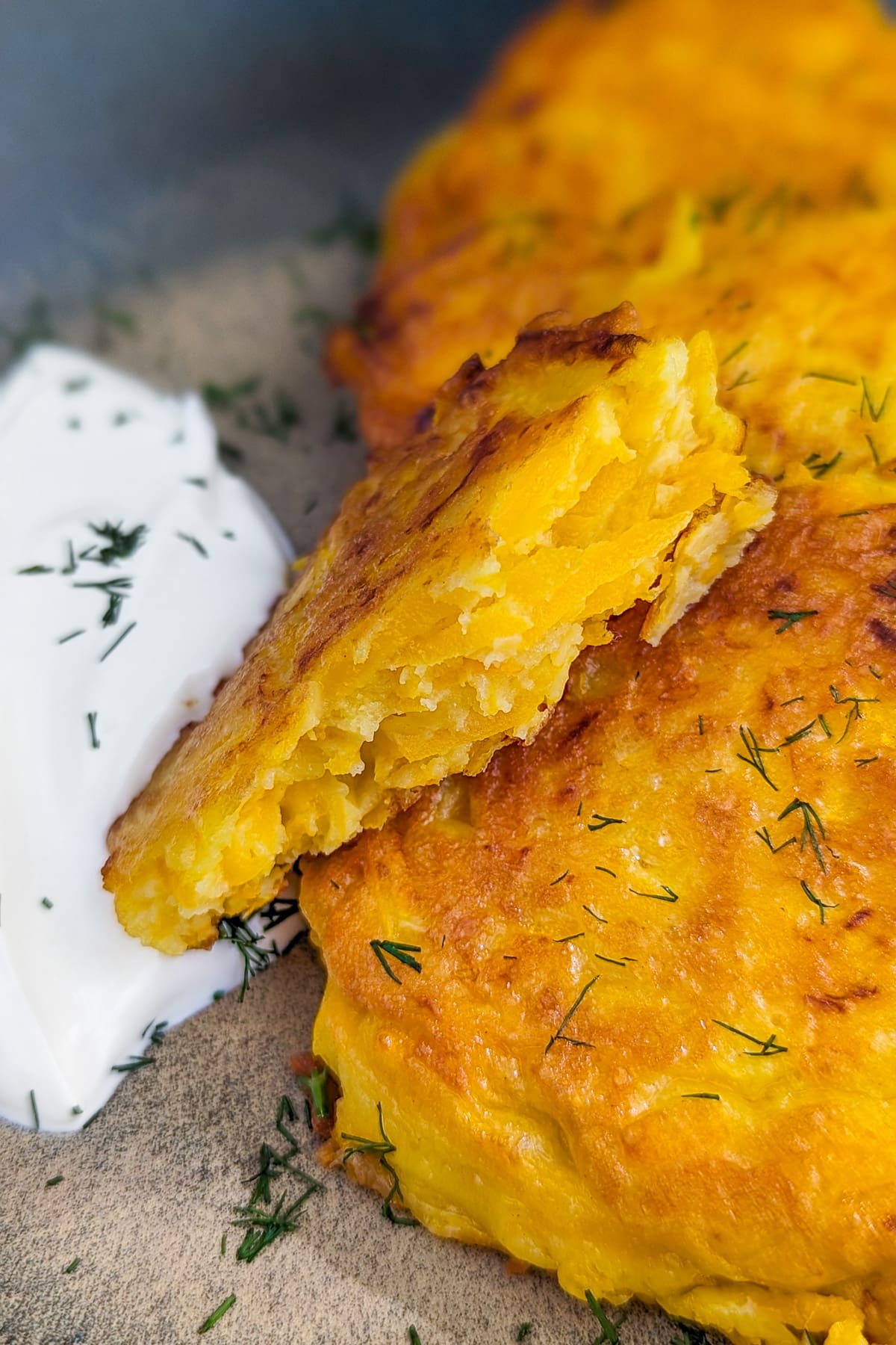 A piece of pumpkin fritters near a stack of fritters served with sour cream and dill.