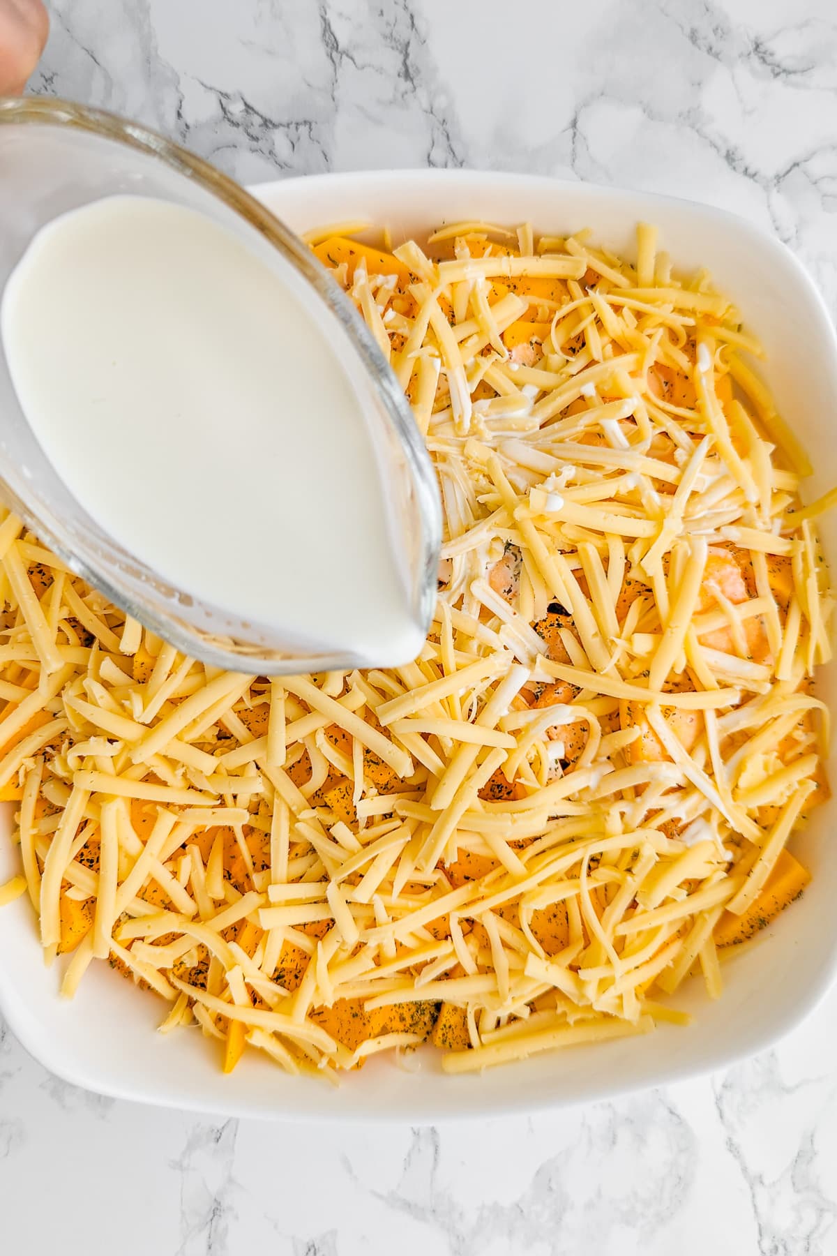 Pouring cream over grated cheese and pumpkin slices in a white casserole.