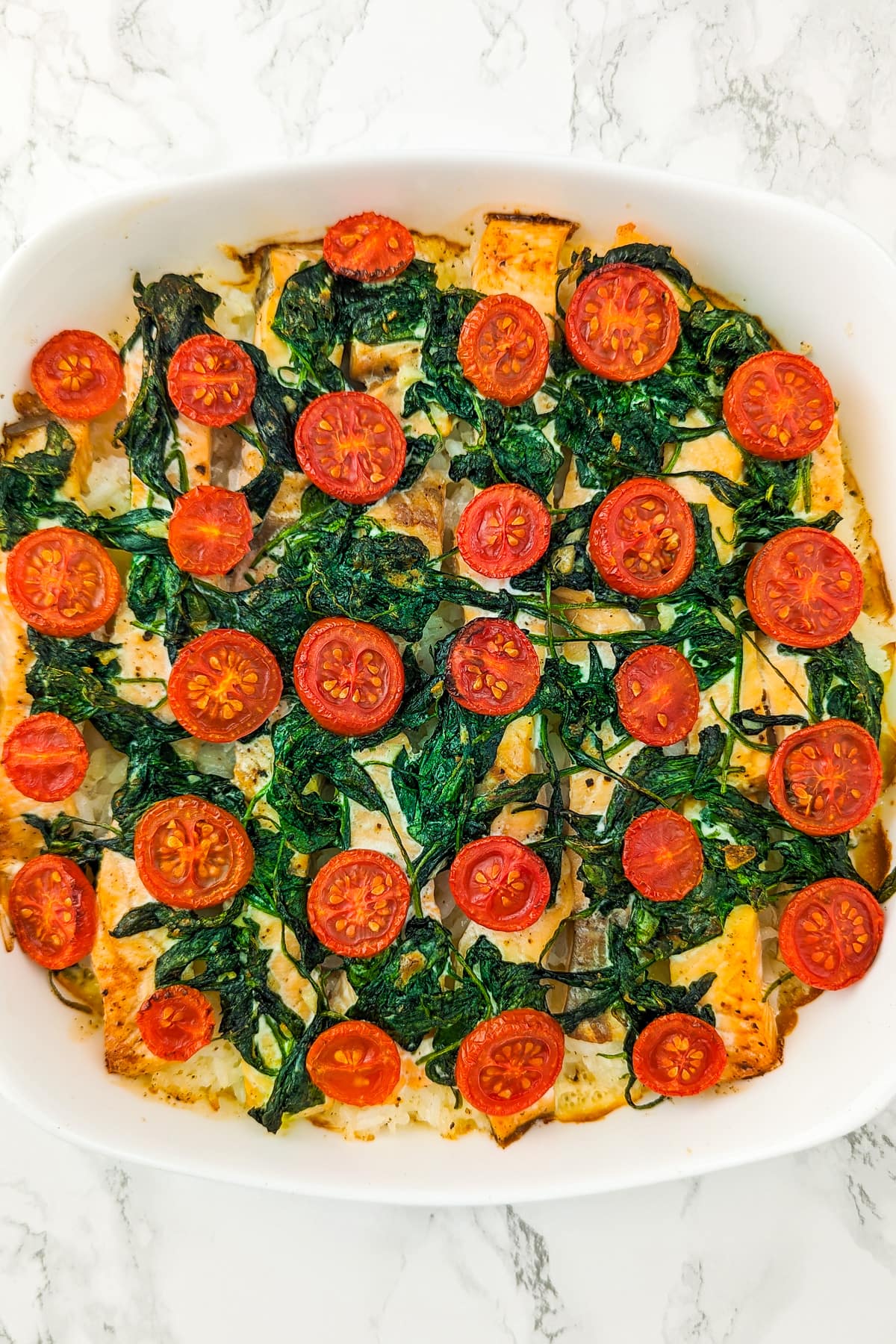 White casserole with boiled rice, salmon slices, cooked spinach, cream and cherry tomato slices.