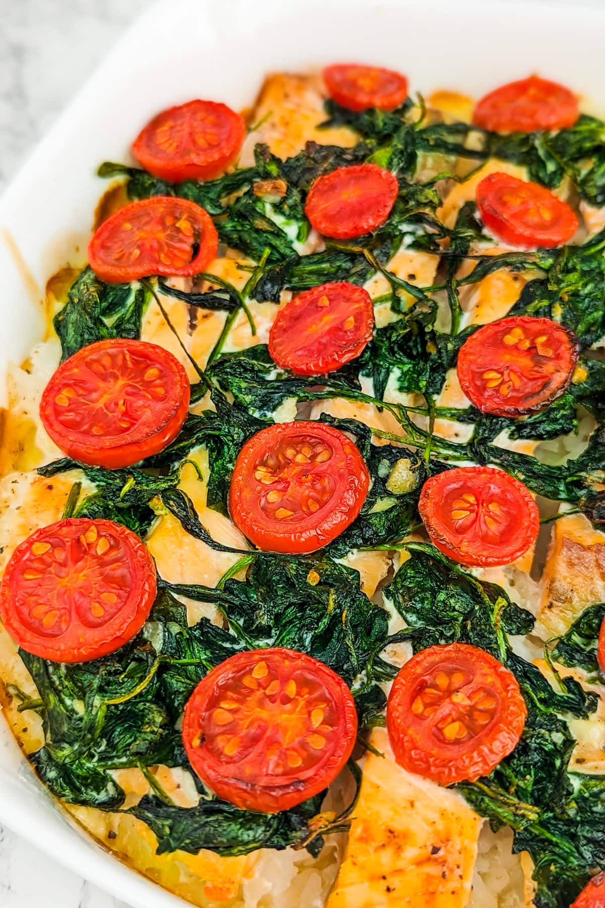 White casserole with boiled rice, salmon slices, cooked spinach, cream and cherry tomato slices.