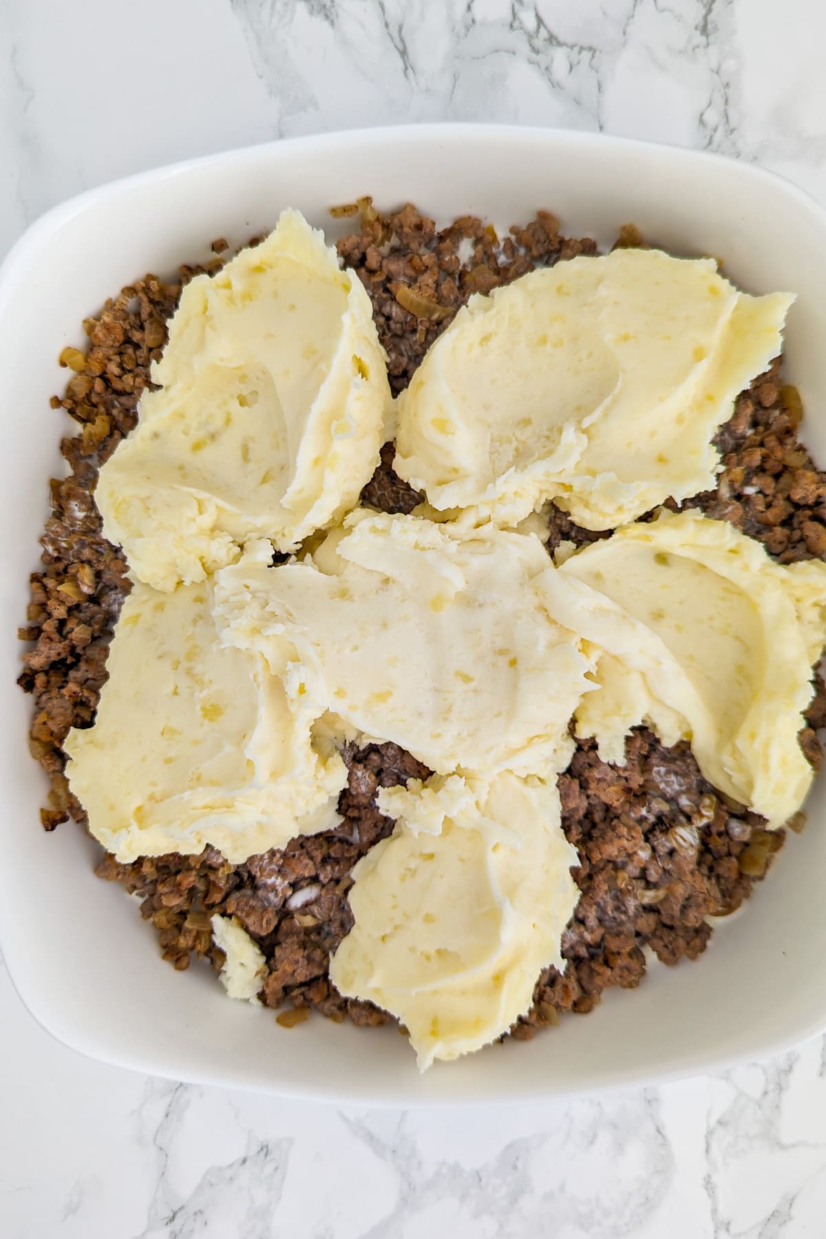 A white casserole with cooked minced meat and covered with a few spoons of mashed potatoes.