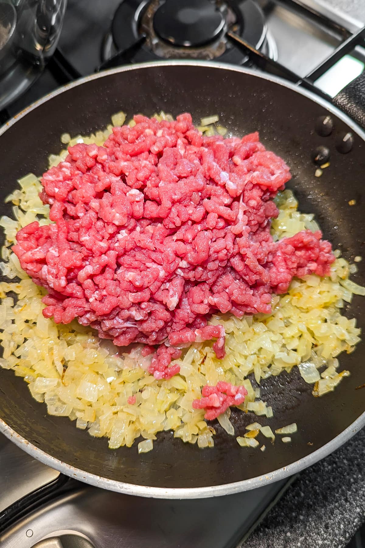 Raw minced meat added over caramelized onions in a frying pan on the stove.
