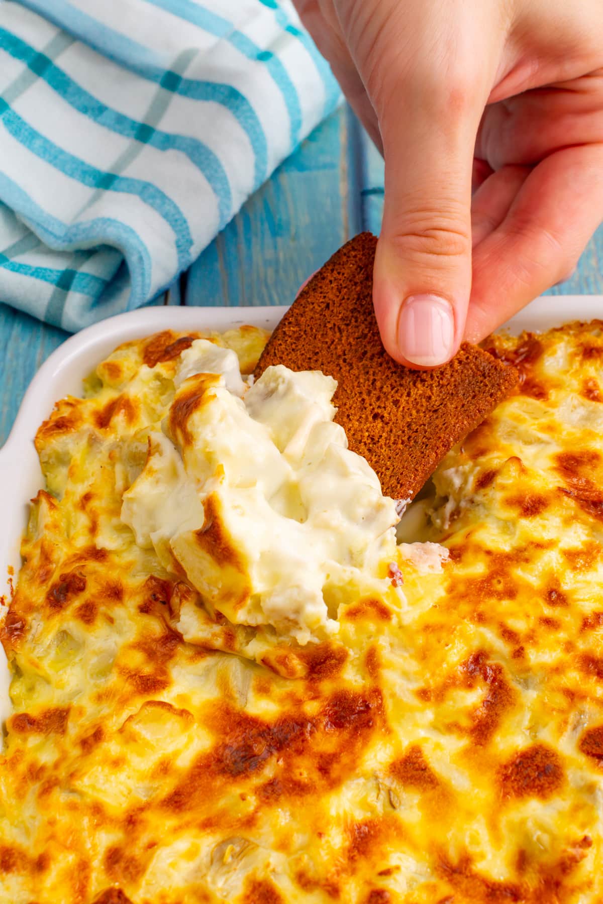 Woman hand holding a crouton with artichoke dip over a white casserole dish with artichoke dip.