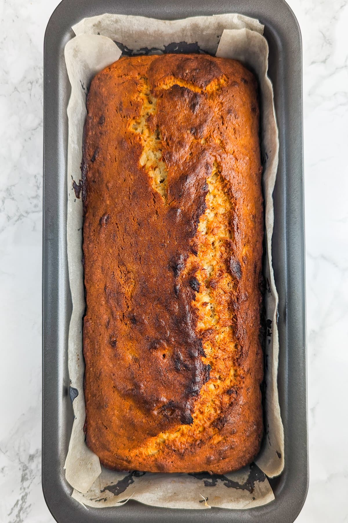 Gray metal cake pan with banana bread inside on a white marble surface.