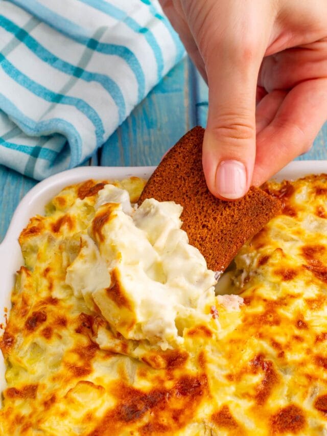 Woman hand holding a crouton with artichoke dip over a white casserole dish with artichoke dip.