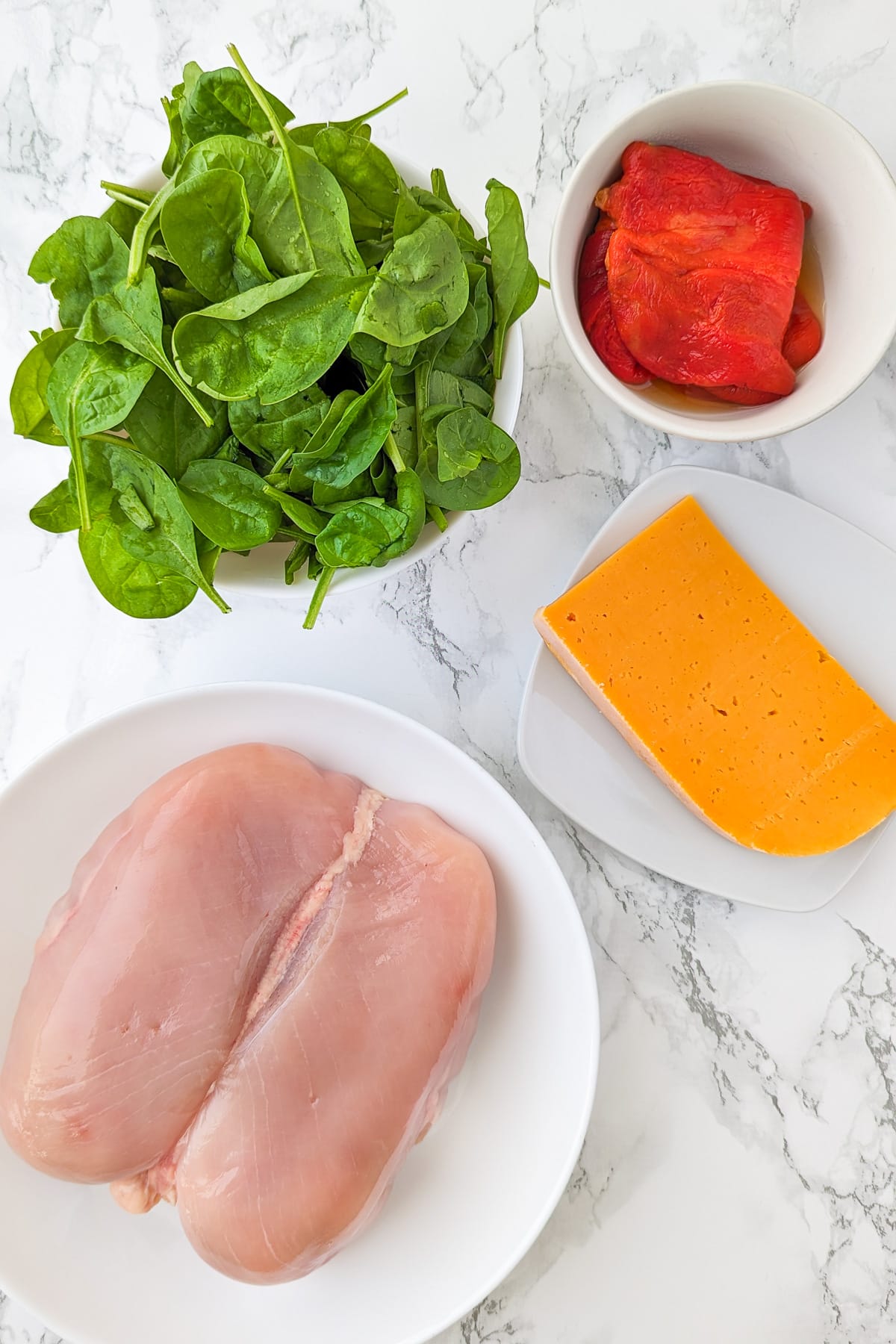 Spinach, roasted bell peppers, cheddar cheese and a chicken breast on a white marble table.