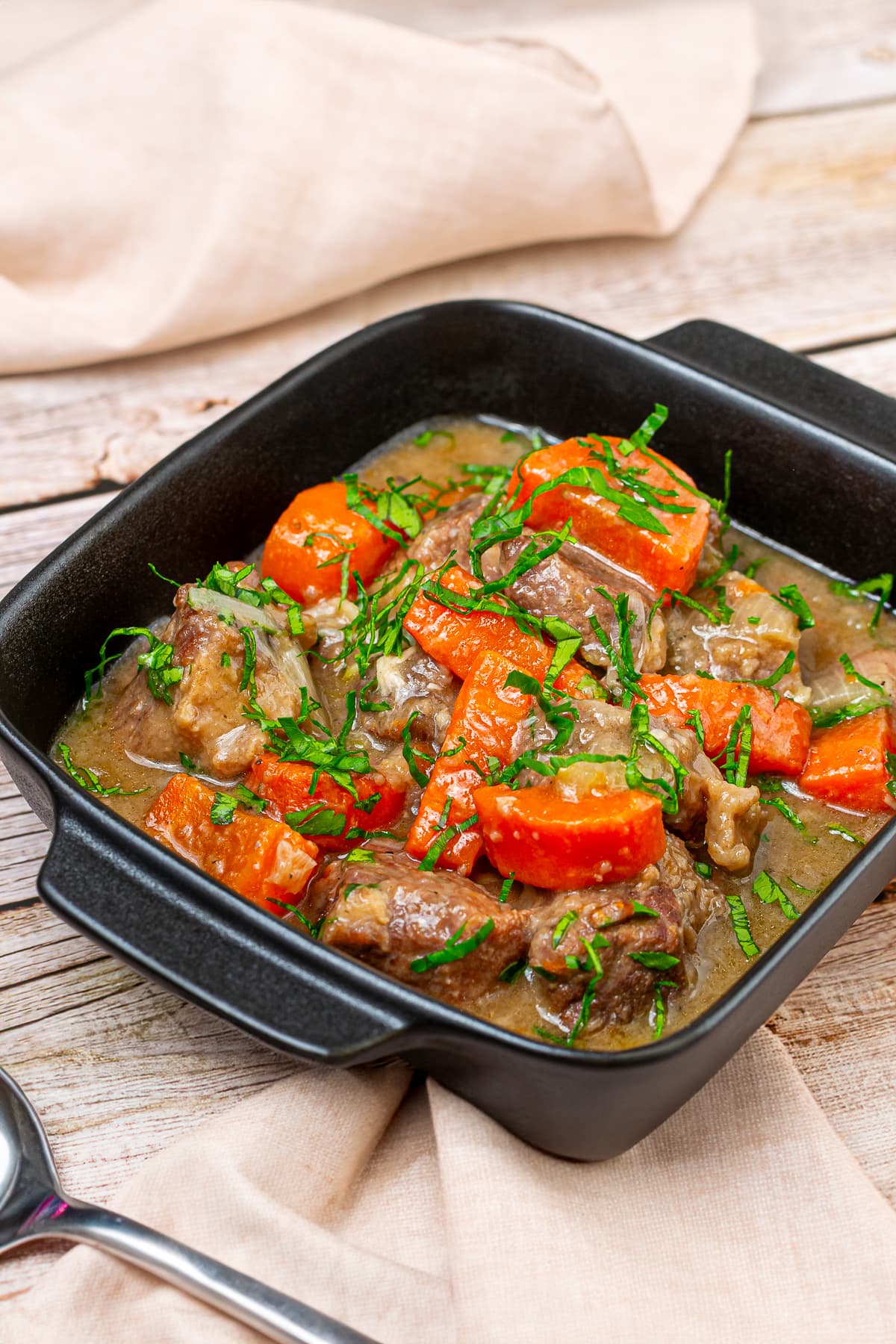 Beef stew with carrots and chopped parsley in a squared black plate on a wooden surface.
