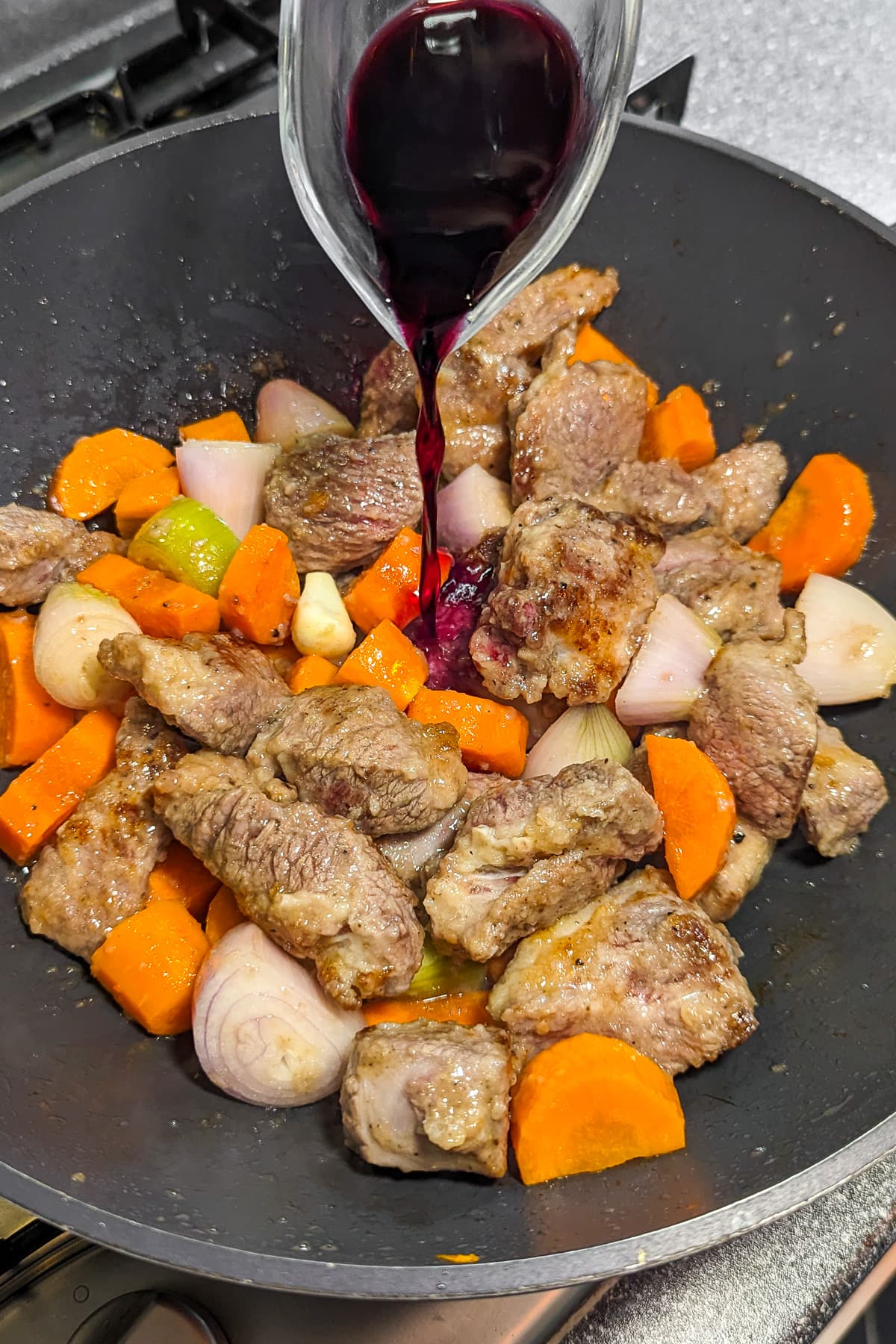 Pouring red wine in a wok with beef, carrots, onions.