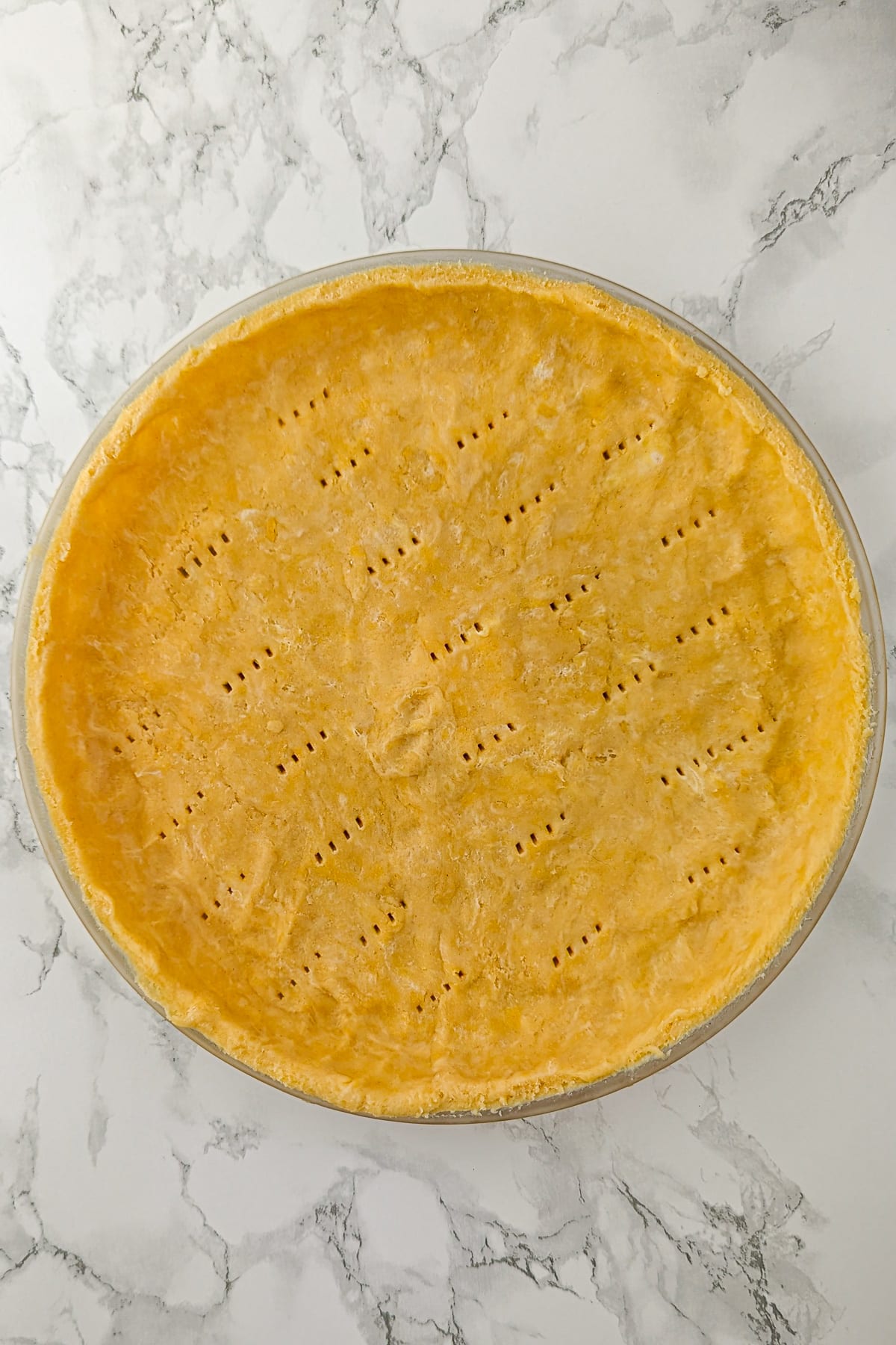 Unbaked homemade quiche dough in a glass pie dish on a marble countertop, with a pattern of fork pricks on the crust indicating it's ready to bake.