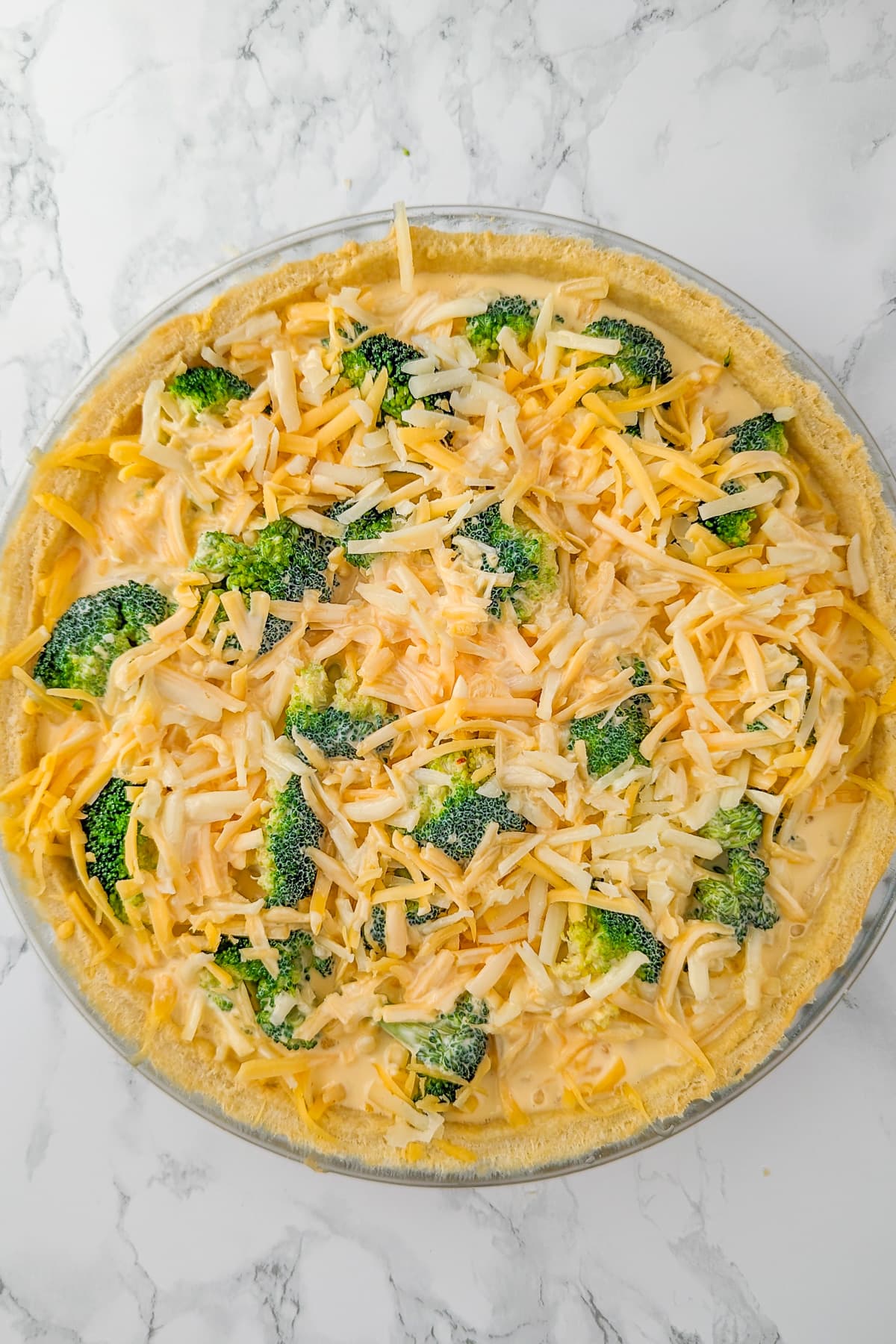 A quiche before baking, where the egg and cream mixture is poured over the broccoli and shredded cheese in the dough crust.