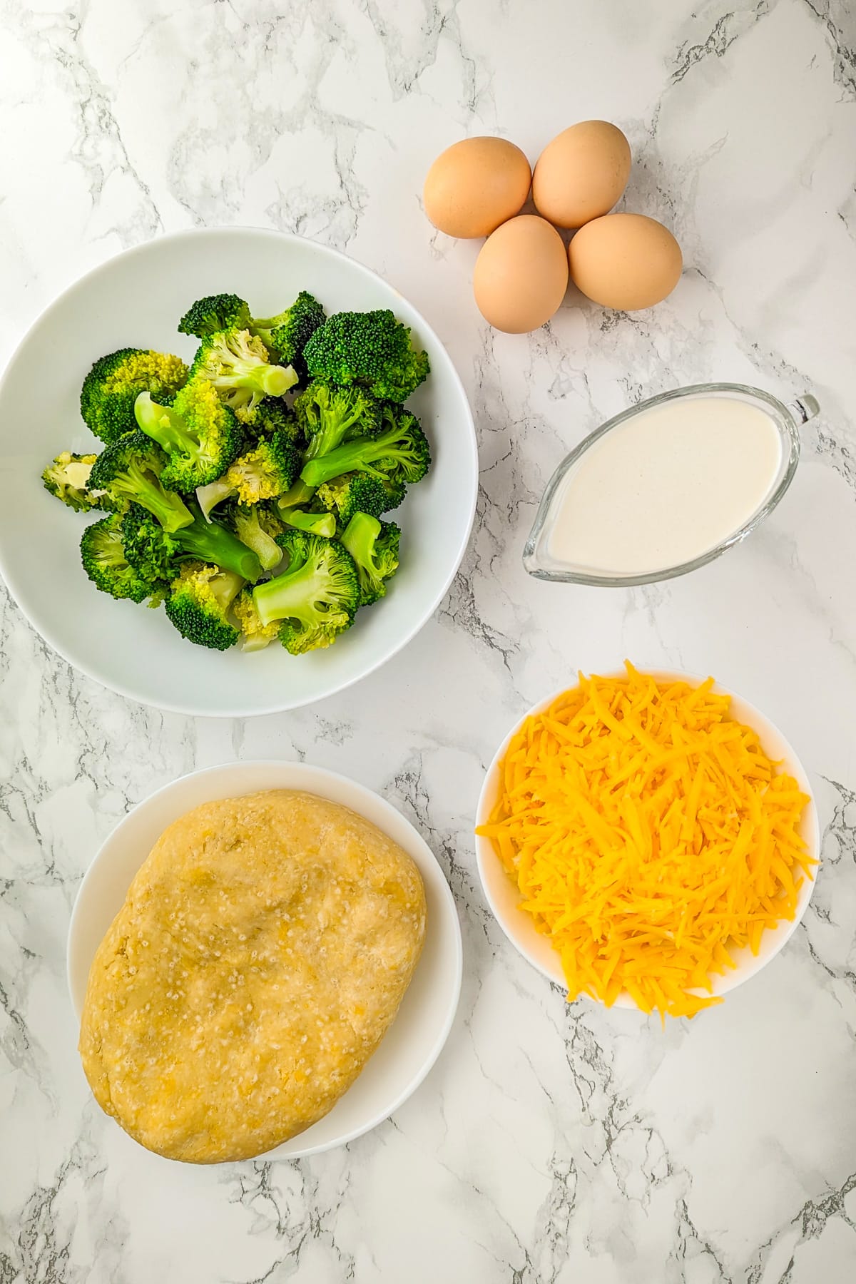 Ingredients for broccoli cheddar quiche on a marble surface, including fresh broccoli florets, shredded cheddar cheese, a dough crust, eggs, and cream.