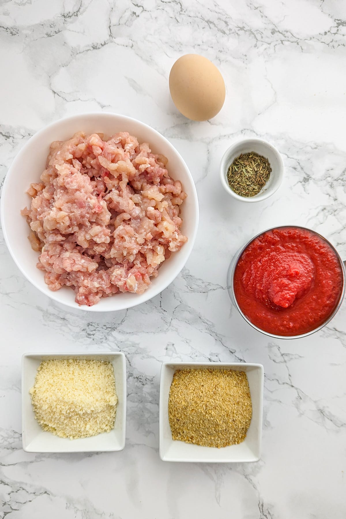 Ground chicken meat, near marinara sauce, parmesan, egg and breadcrumbs on a white marble surface.