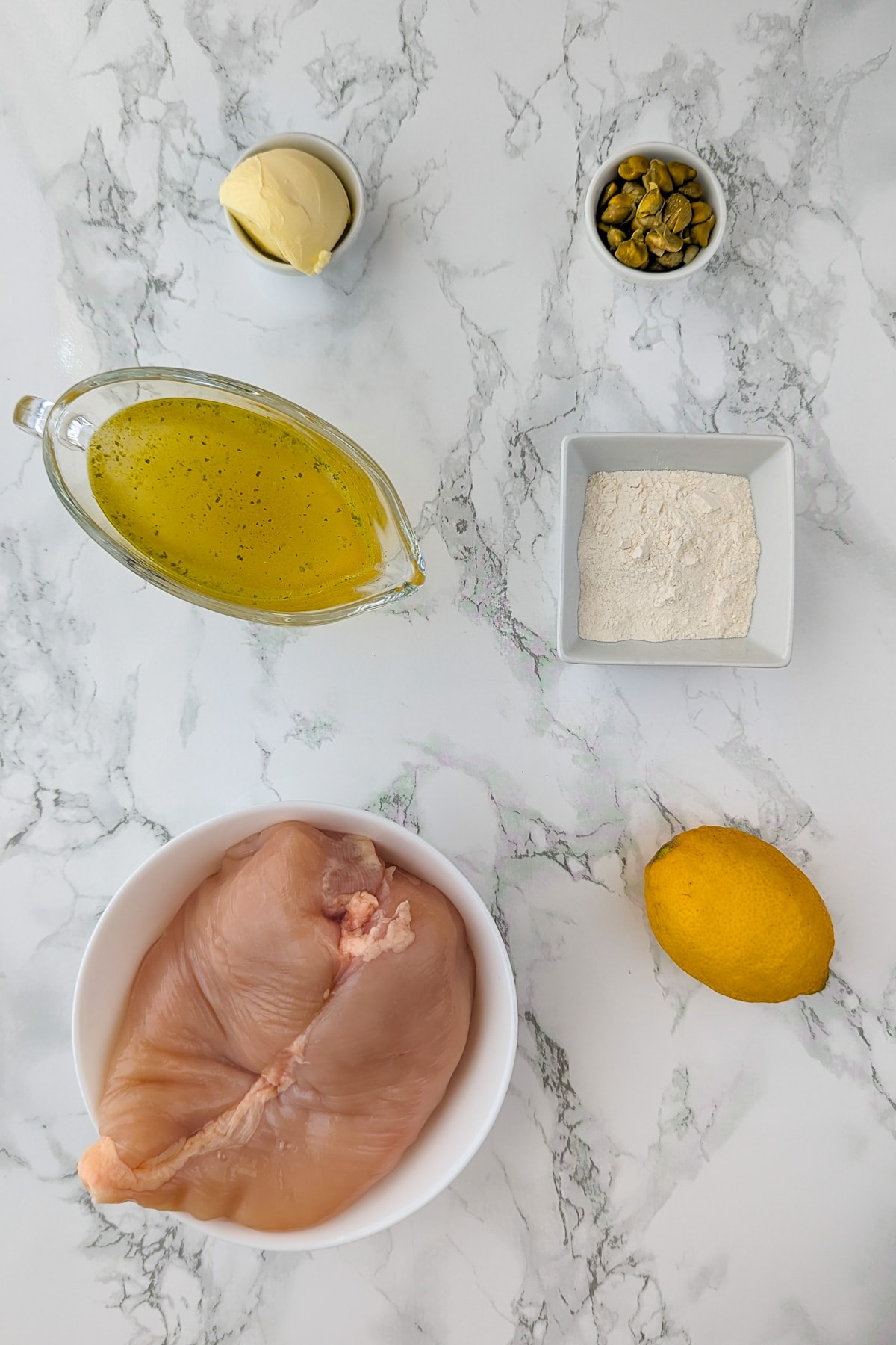 Ingredients for chicken piccata neatly arranged on a marble countertop, including raw chicken breasts, lemon, capers, butter, flour, and a jug of broth.