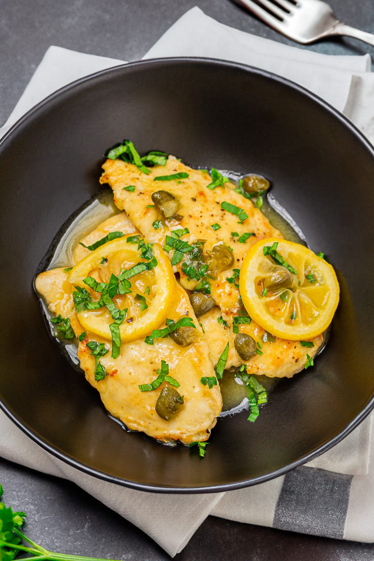 Plate of chicken piccata garnished with sliced lemon and capers on a dark table, capturing the vibrant colors and texture of the dish.