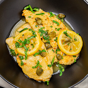 A vibrant dish of chicken piccata, featuring golden-browned chicken breasts topped with lemon slices and capers, garnished with chopped parsley, presented in a dark bowl on a grey background.